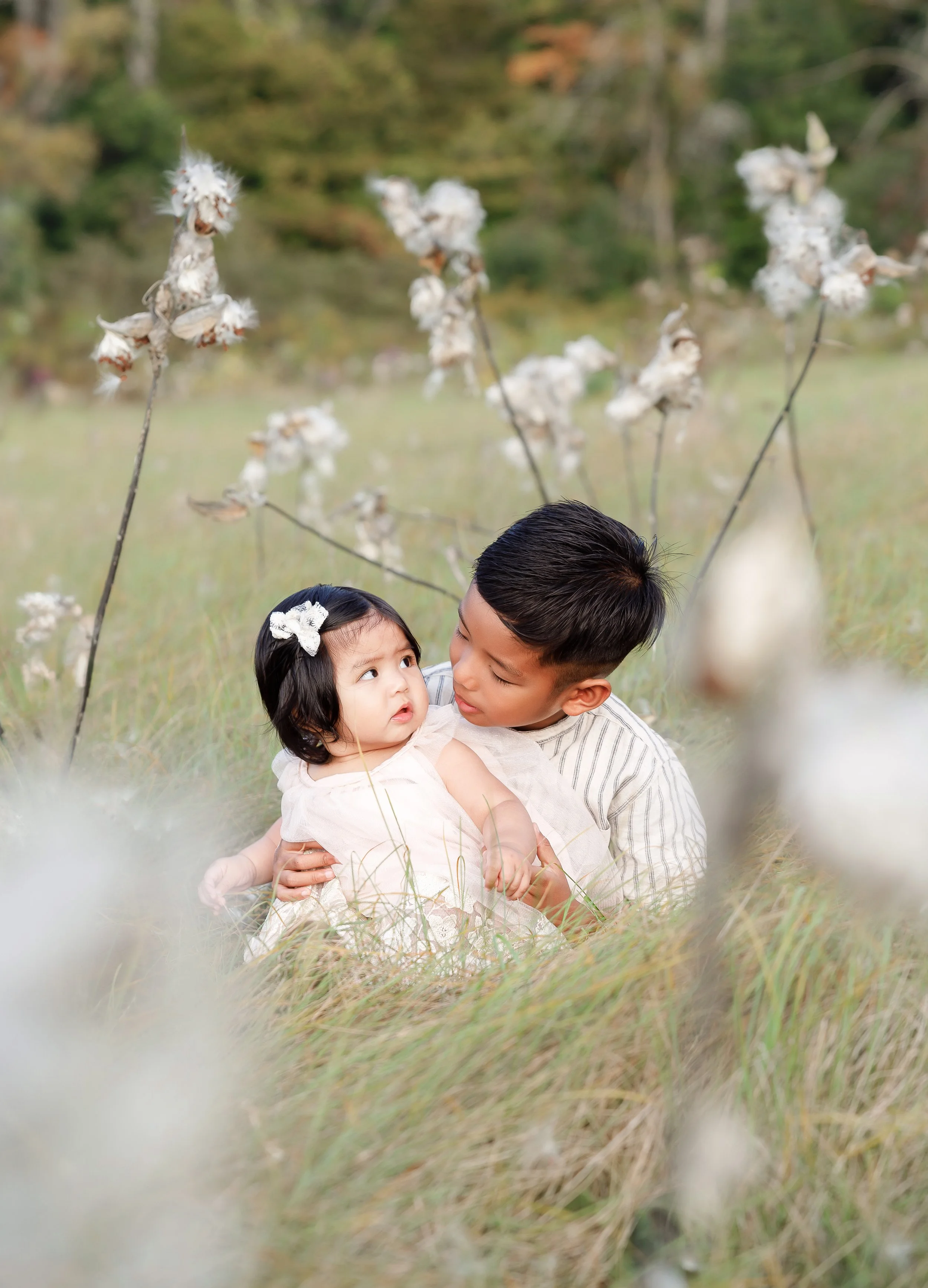A young man and a small girl sitting together in a grassy field during fall, surrounded by cotton plants and autumn trees in the background.
