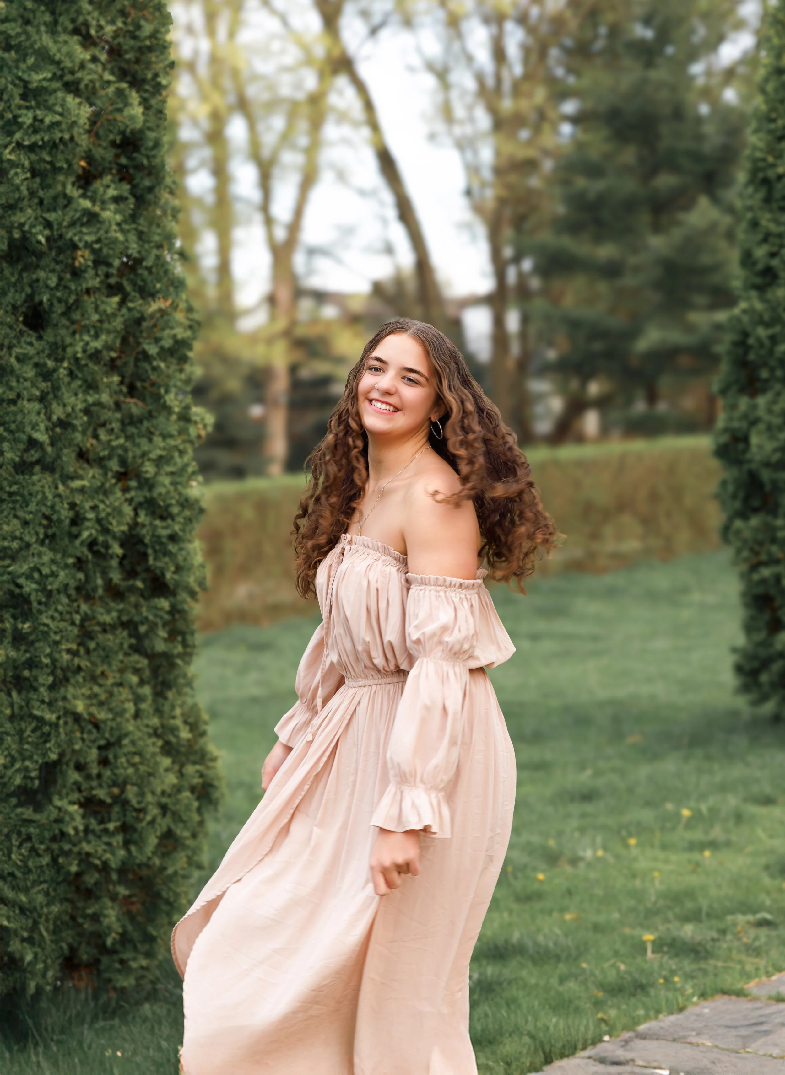 Young woman with curly hair and a light pink off-shoulder dress standing outdoors in a park with greenery and trees in the background.