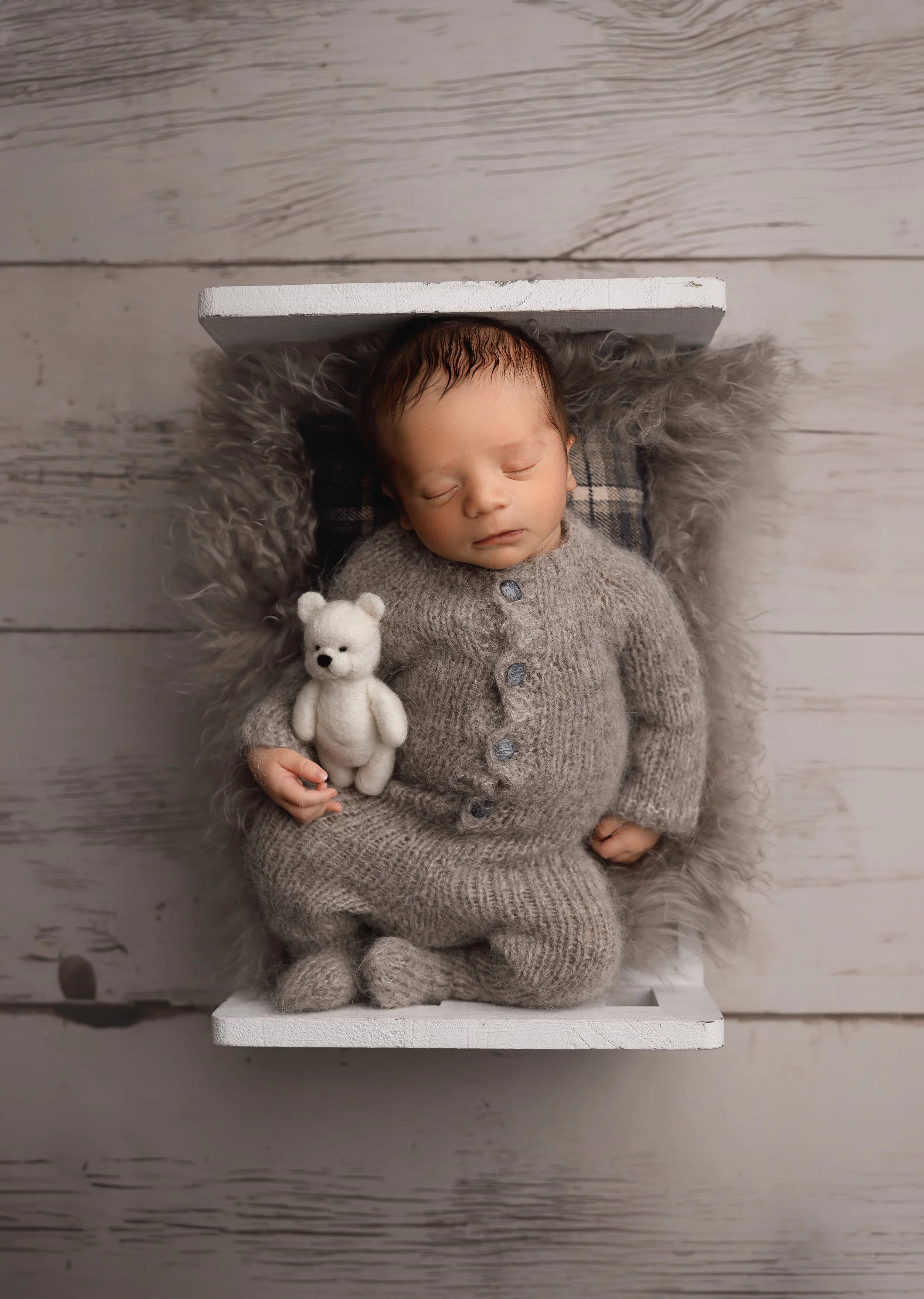 A newborn baby girl sleeping on a soft, furry gray blanket with a plaid pillow behind her head, dressed in a gray knitted sweater with a bear plush toy on her chest, on a white wooden platform.