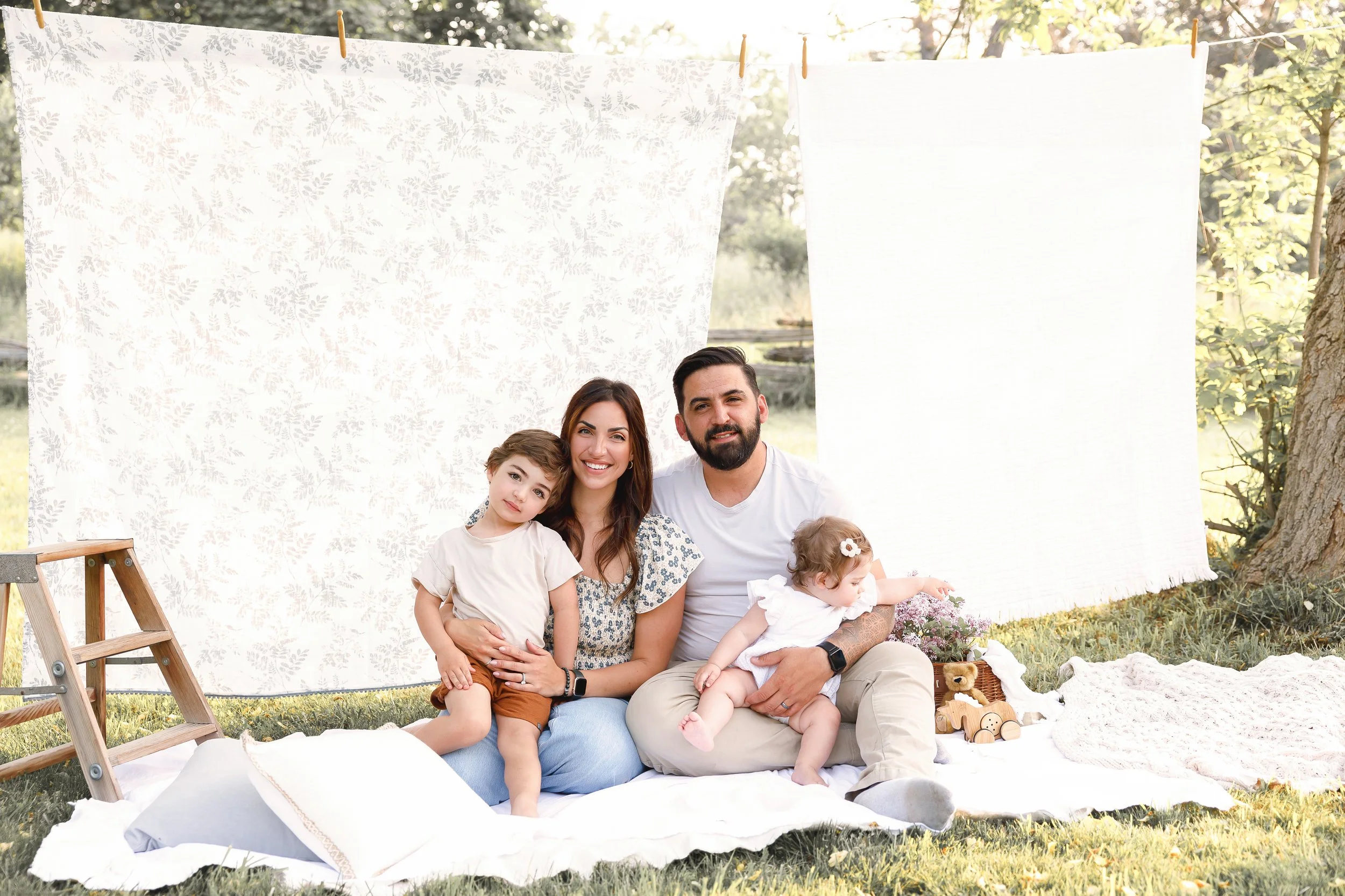 Family of five sitting outdoors in a backyard, smiling, with a white patterned blanket hanging on a clothesline behind them, surrounded by trees and flowers, enjoying a sunny day.