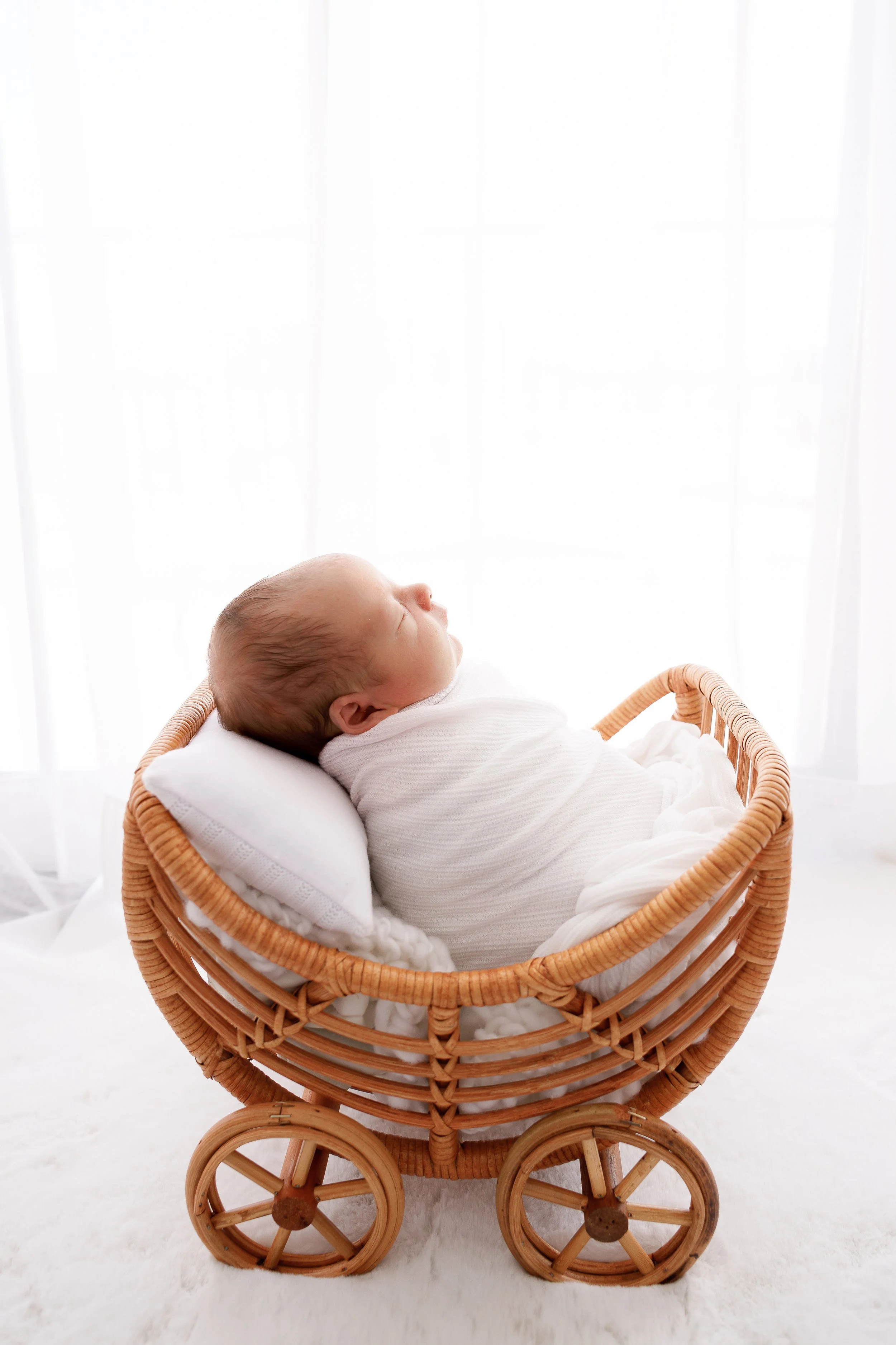 A sleeping baby in a white outfit lying in a wicker doll carriage with a white pillow in a bright, softly lit room.