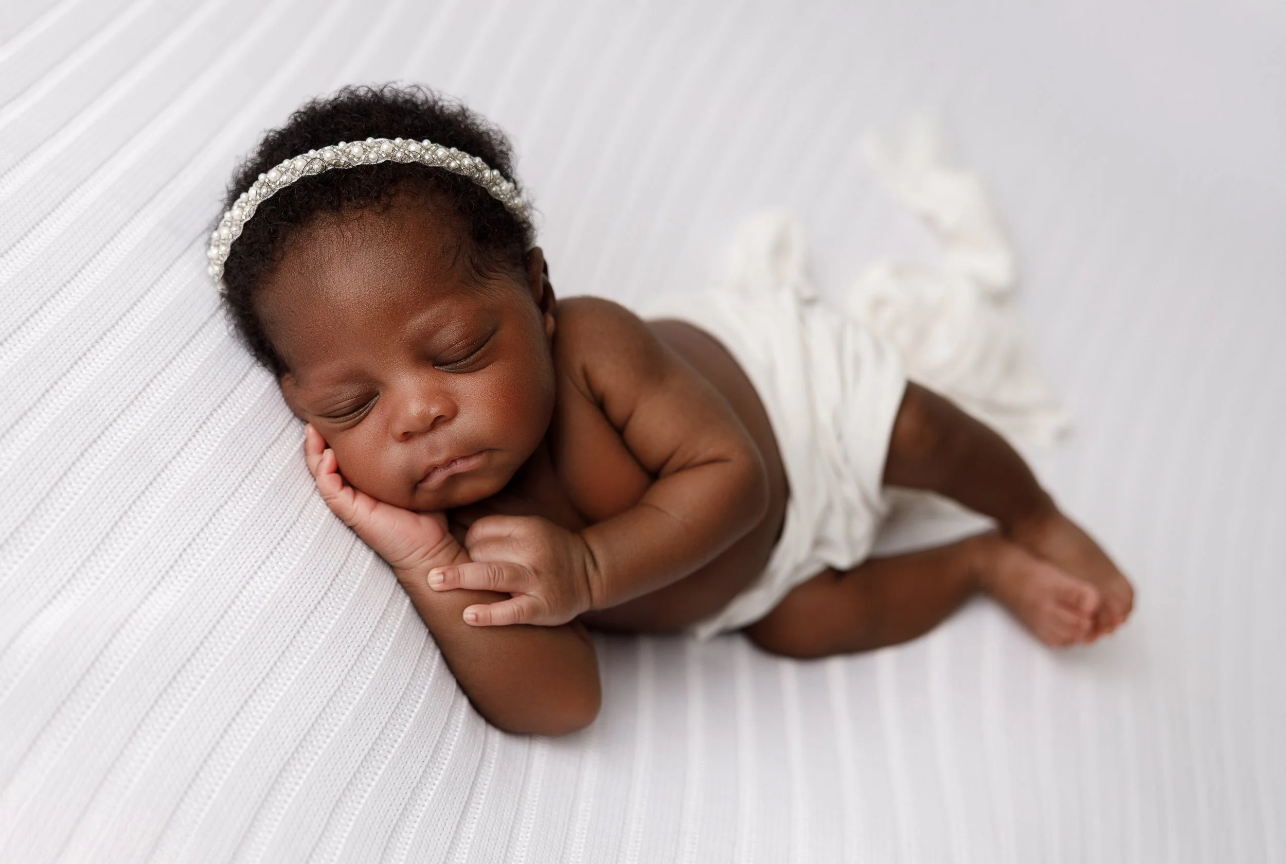 A sleeping baby girl with curly hair wearing a pearl headband, resting on a white textured surface.