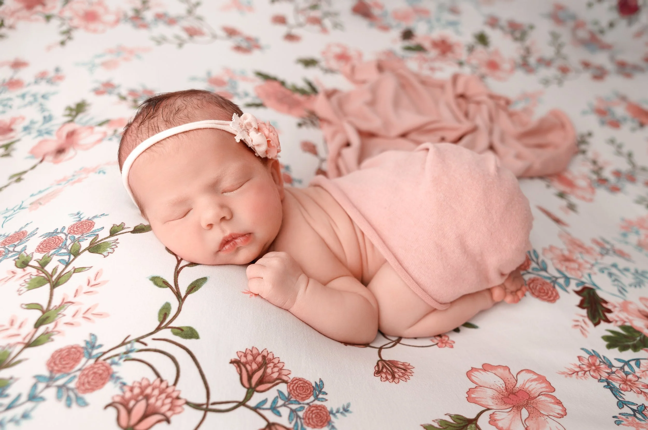 A sleeping newborn baby with a pink headband and floral decoration, lying on a floral patterned fabric with pink, green, and blue colors.