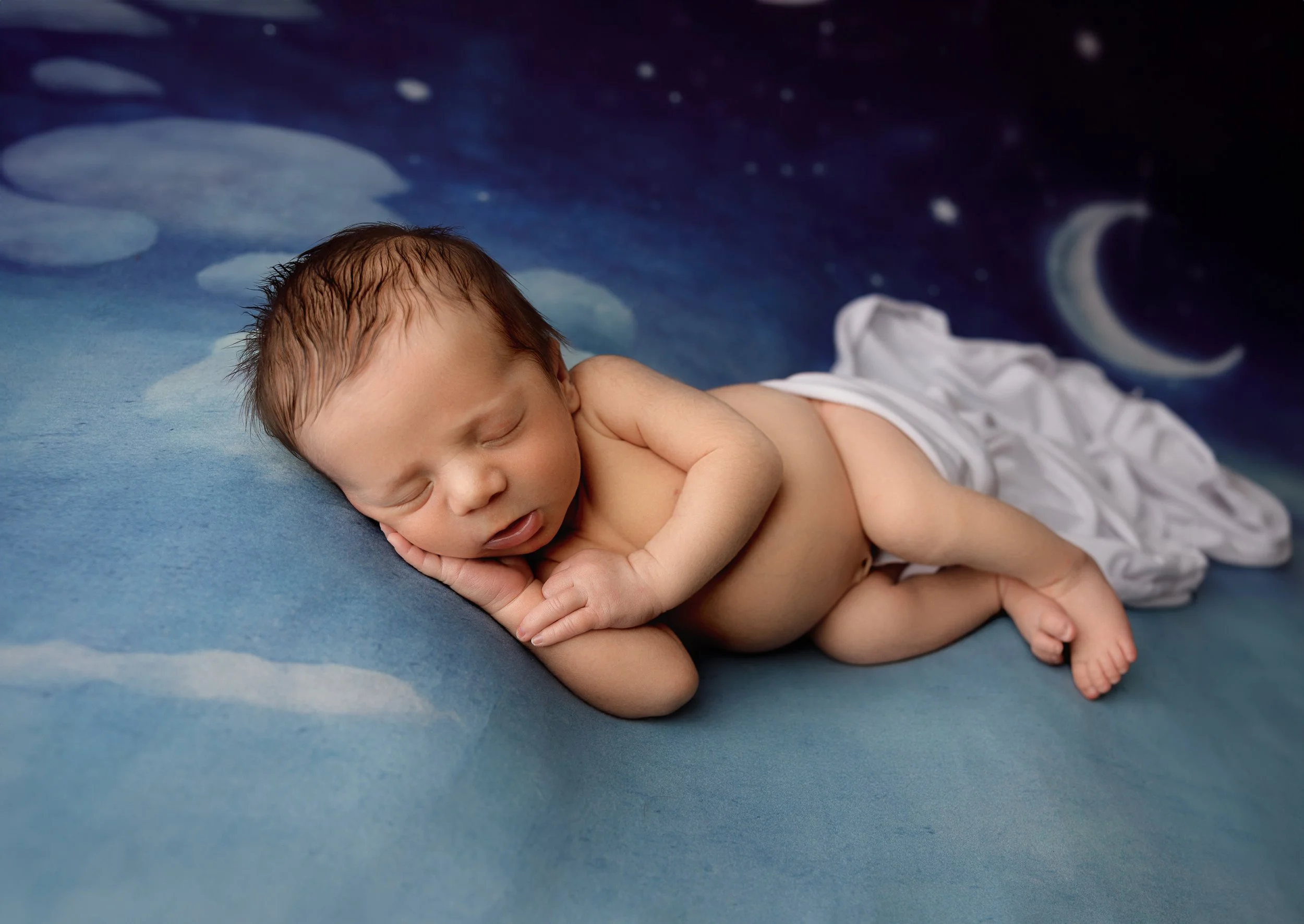 A sleeping baby with light brown hair resting on a blue surface with a space-themed background, including a moon, stars, clouds, and a crescent moon.