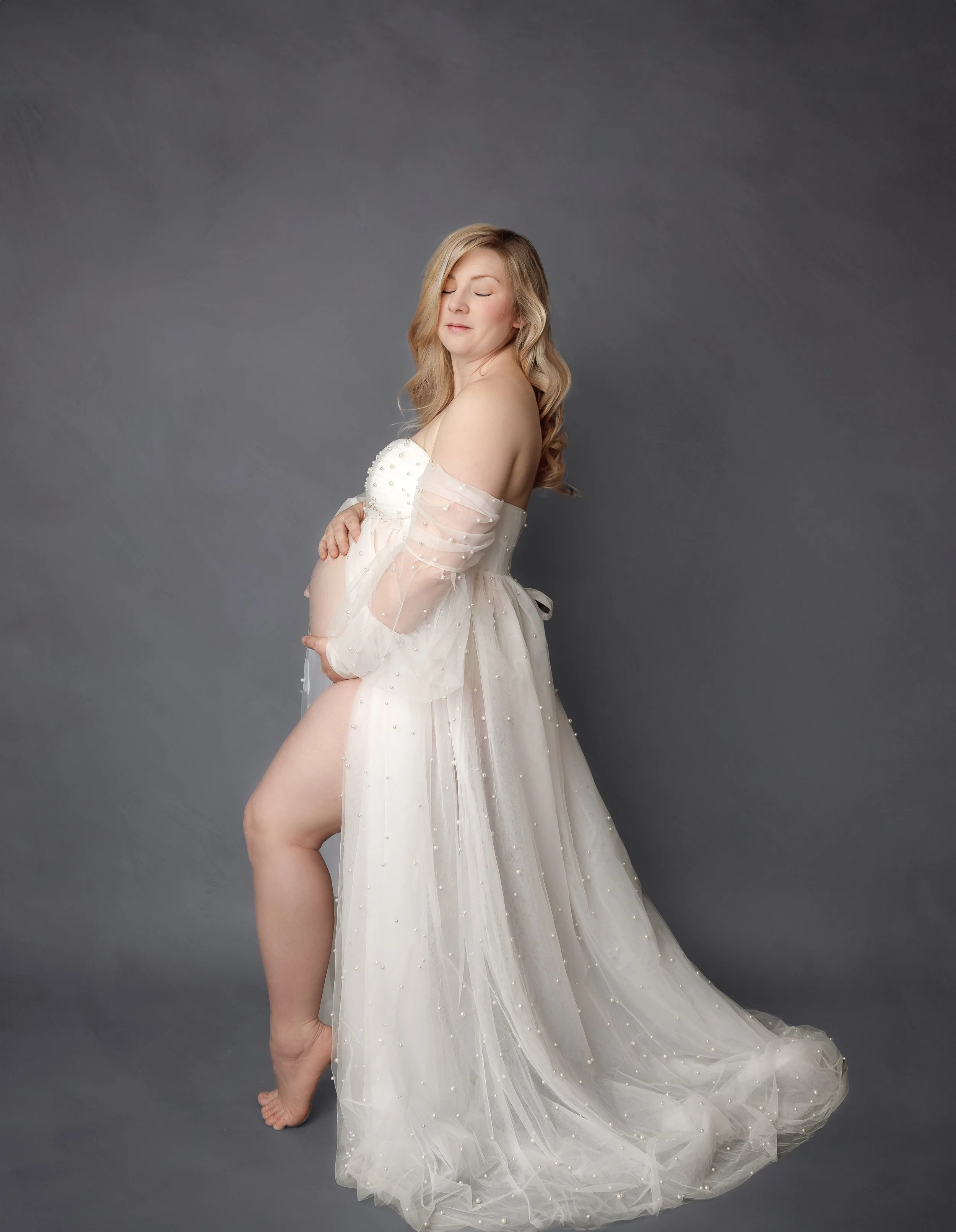 A pregnant woman with blonde hair in a wedding dress with off-the-shoulder sleeves, standing against a gray background. She has her eyes closed, cradling her belly with both hands, and looks peaceful.