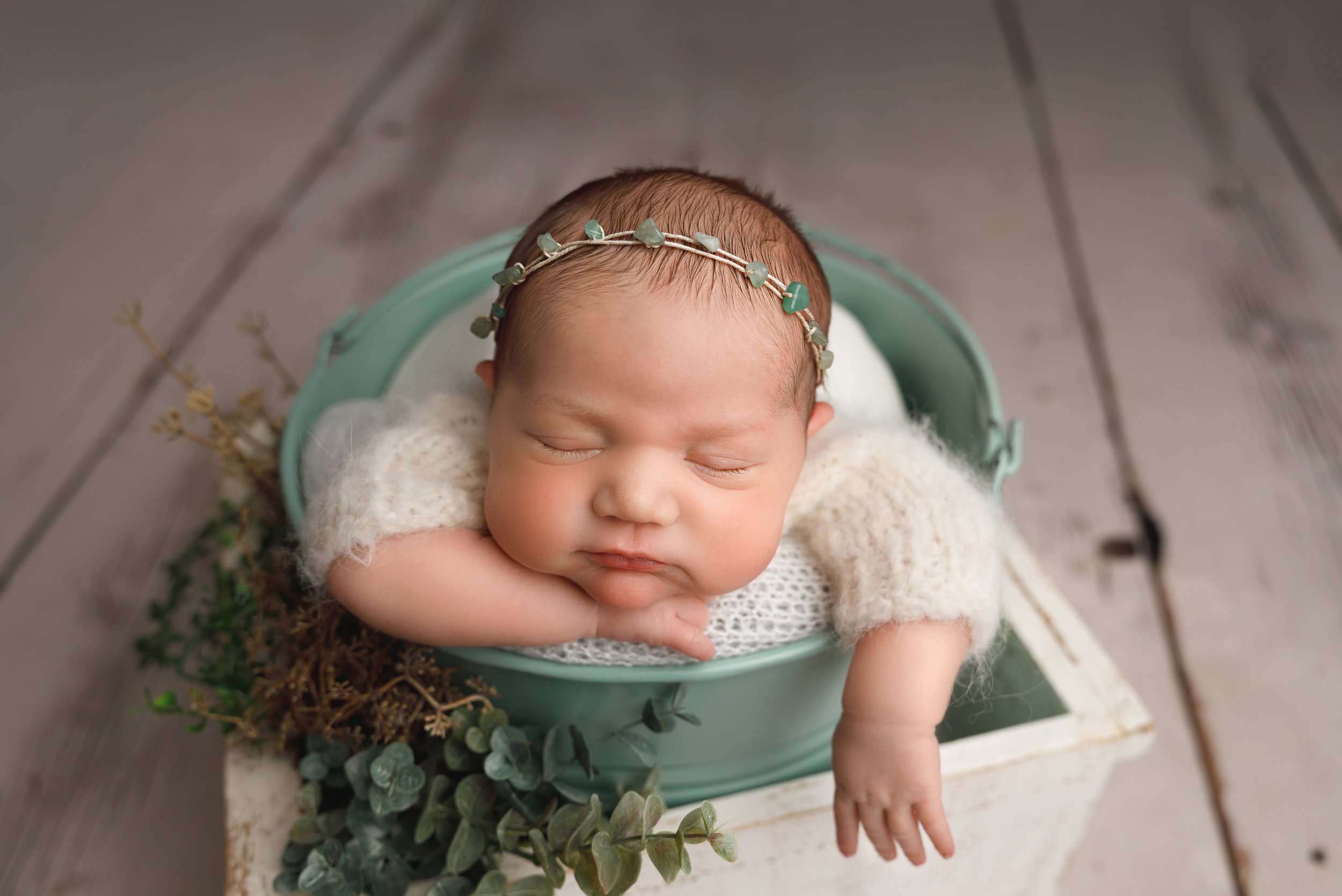 A sleeping baby with a floral headband lying in a green bucket lined with white cloth, surrounded by decorative greenery on a wooden surface.