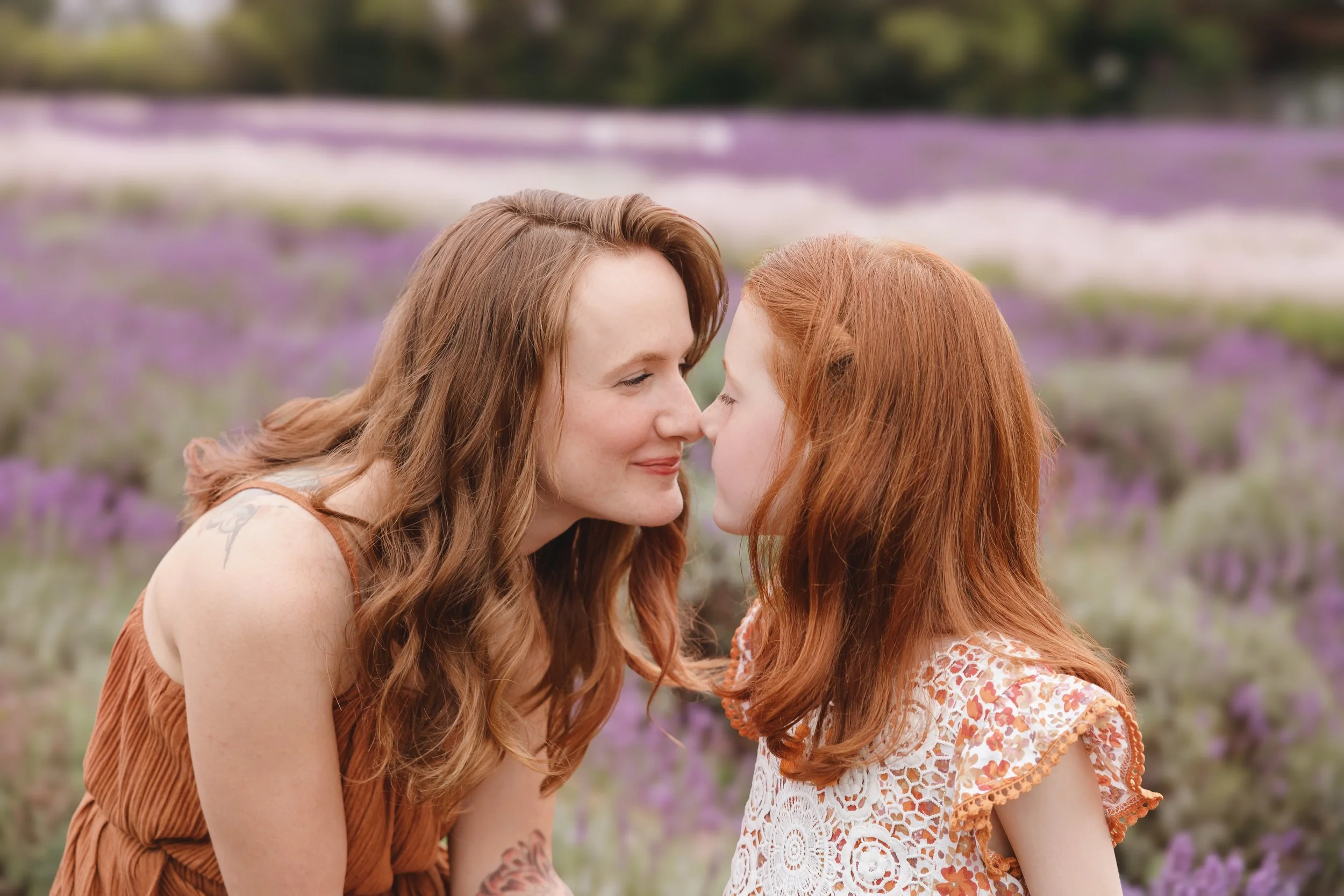 A woman and a young girl with red hair sharing a close moment outdoors with purple flowers in the background.