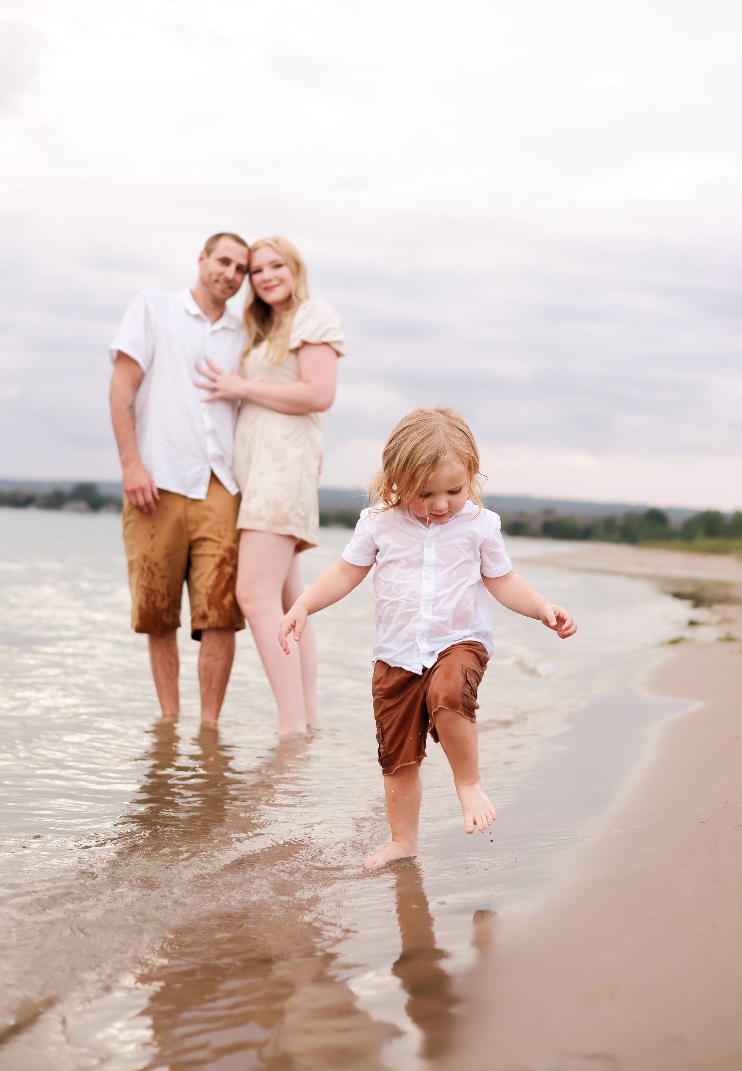 A family of three walking along the shoreline at the beach. The father and mother are smiling and holding hands, while their young daughter, barefoot and looking down, is walking in front of them.