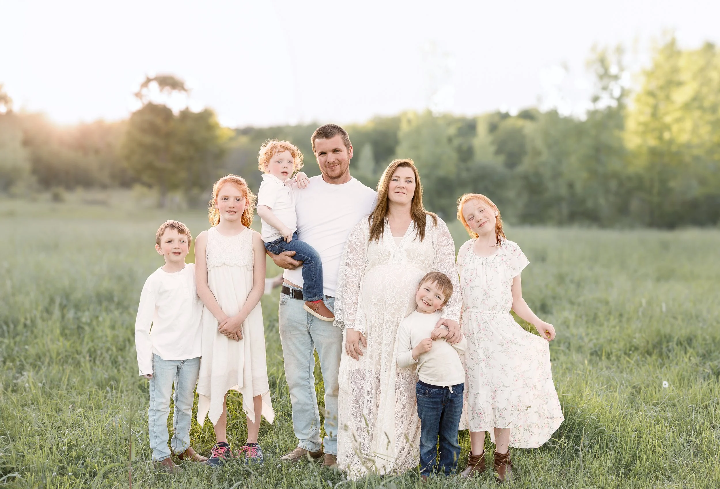 A multi-generational family standing outdoors in a grassy field with trees in the background, basking in sunlight.
