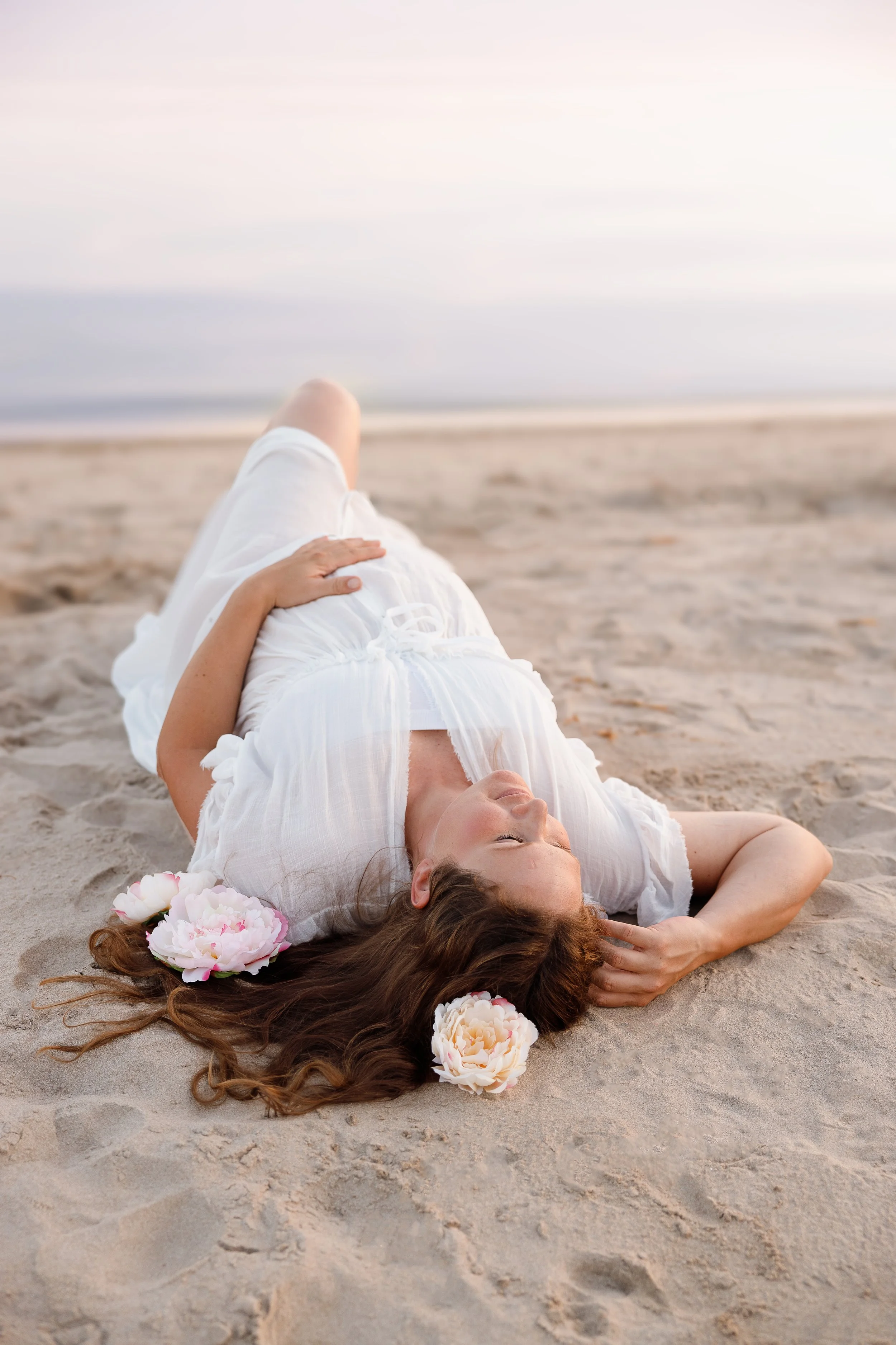 A woman lying on the sandy beach with flowers in her hair, wearing a white dress, relaxing with a view of the ocean and sky in the background.