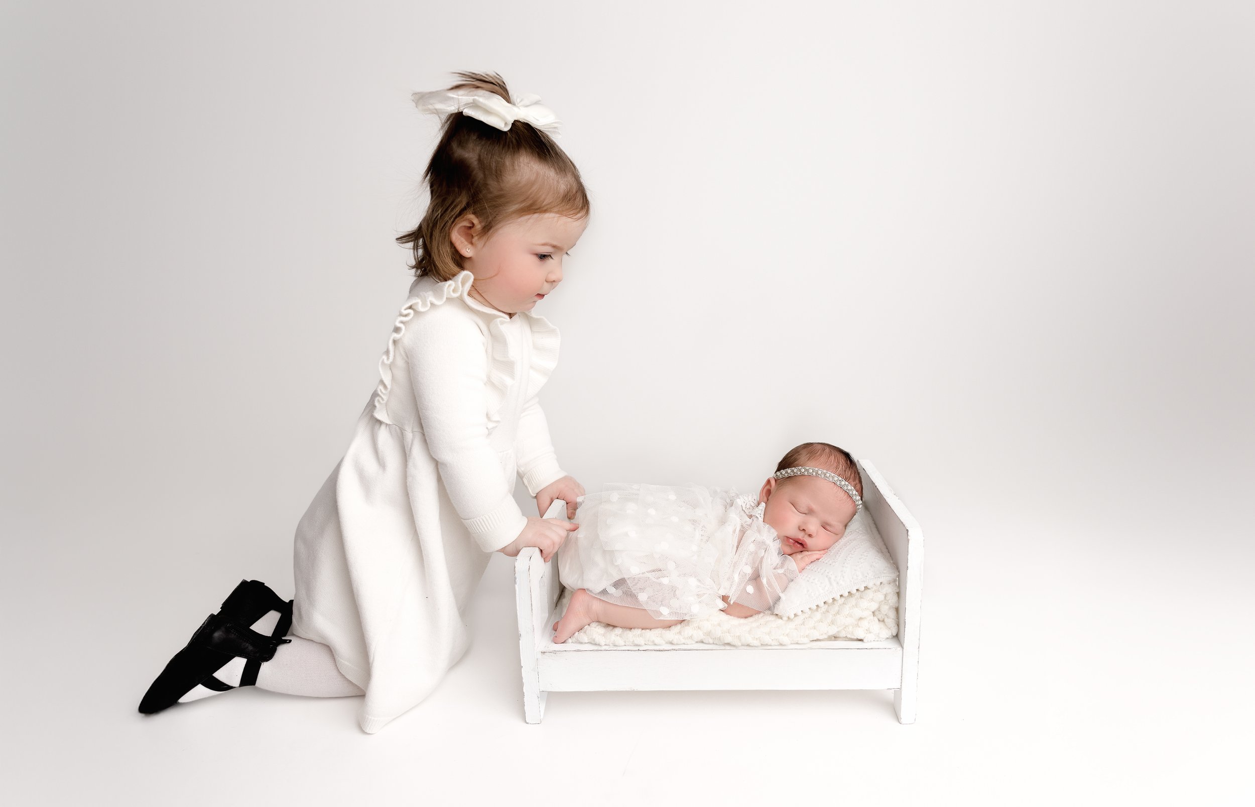A young girl with a white bow in her hair, dressed in white, kneeling beside a baby girl sleeping in a white crib, wearing a lace headband and a pink dress with lace, against a plain white background.