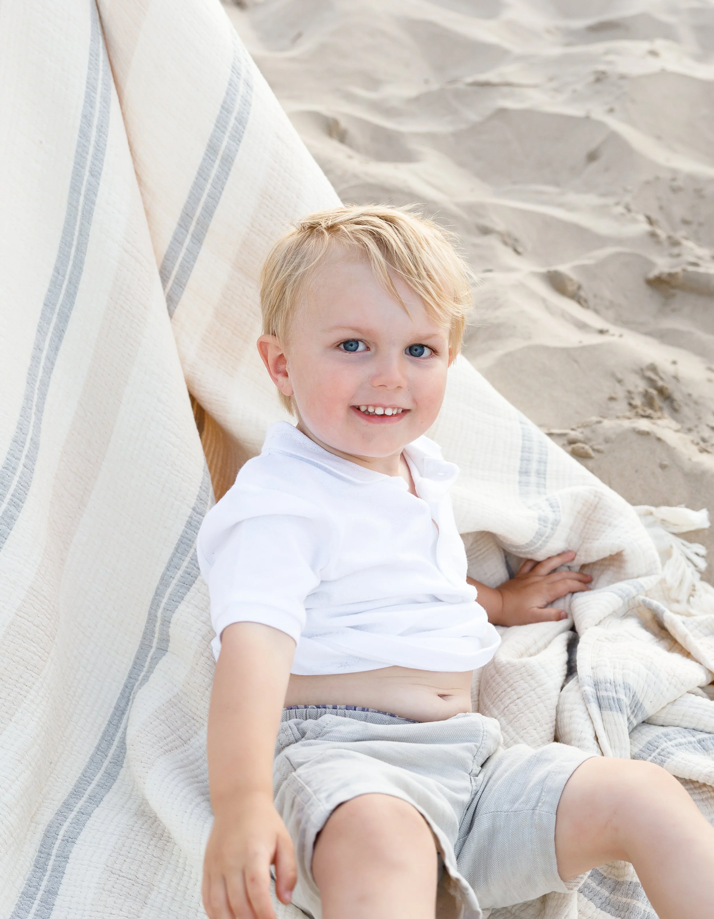 A young boy with blonde hair and blue eyes sitting on a beige blanket on a sandy surface, smiling and looking at the camera.
