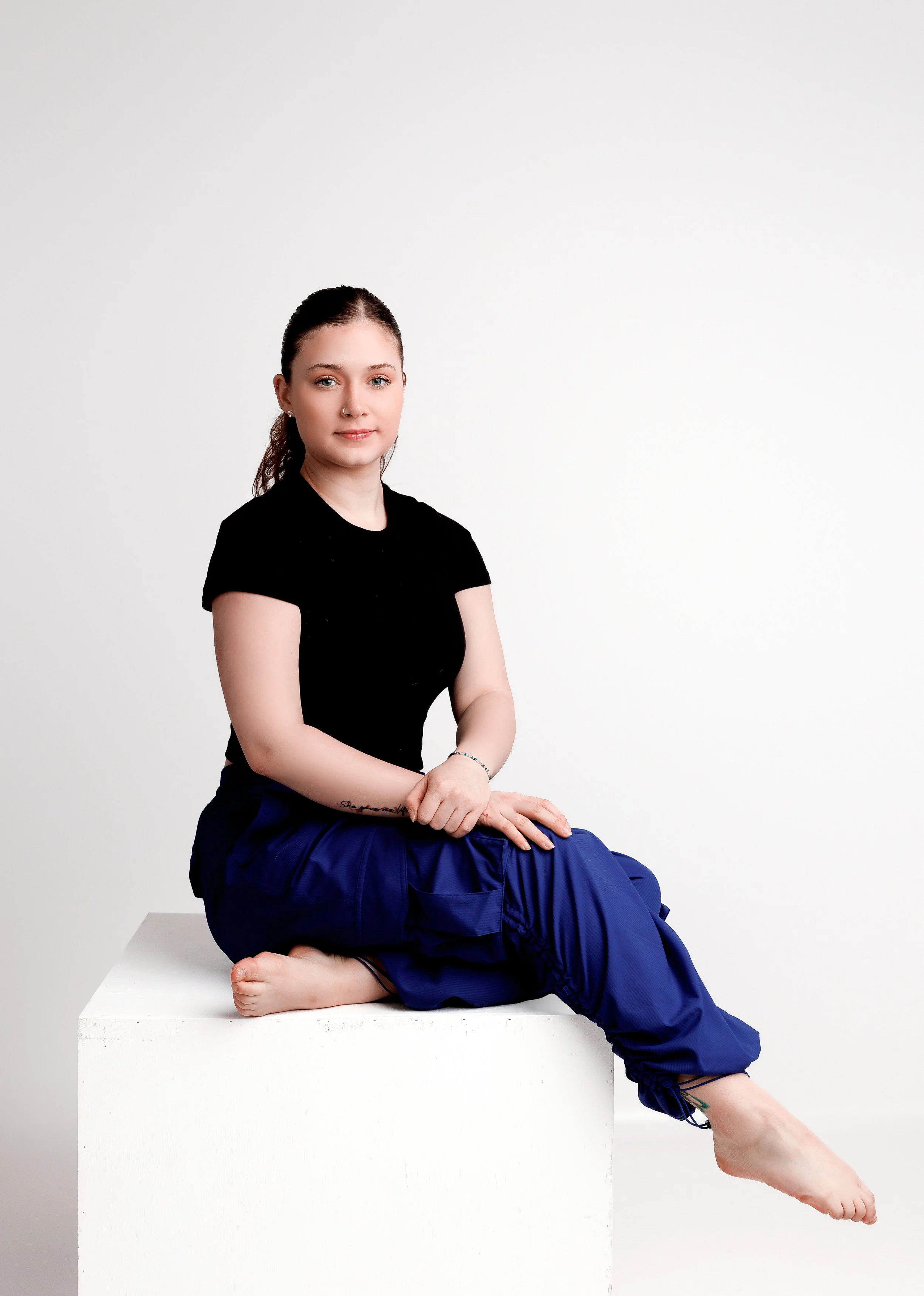 A woman with dark hair, sitting on a white block, wearing a black top and blue pants, posed with her body twisted and her left leg extended. The background is plain white.