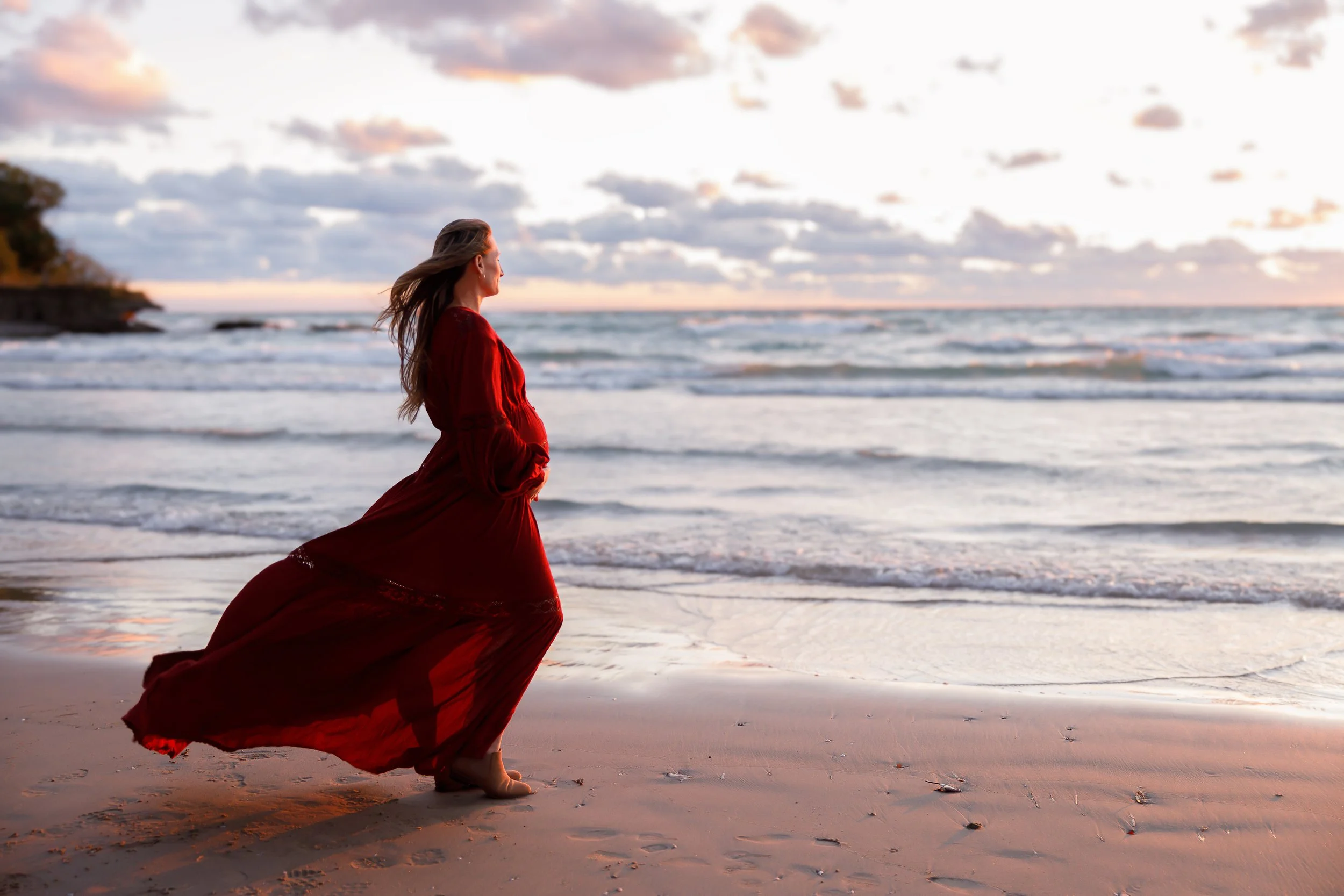 A woman in a flowing red dress walking barefoot on the beach at sunset, with her hair flowing in the breeze. The sky is partly cloudy with a mix of pink and orange hues near the horizon.