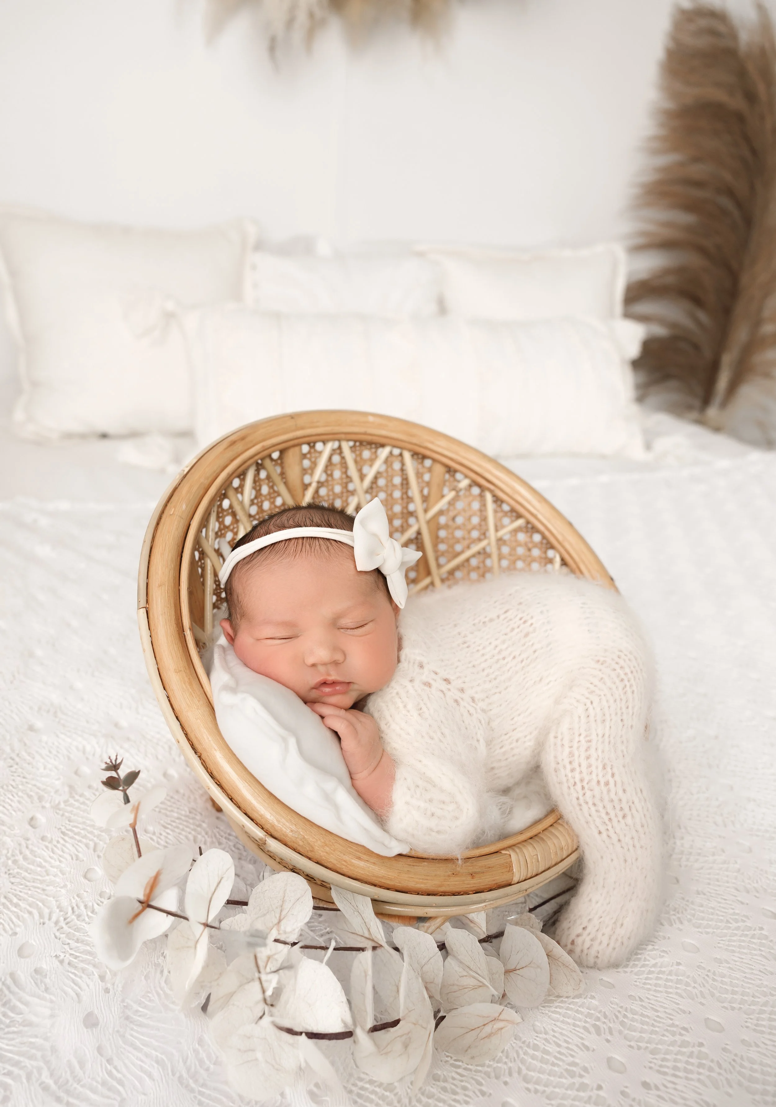 A baby with a white bow headband sleeping in a wicker bassinet on a white lace blanket, with dried leaves and flowers nearby, in a bright, soft setting.