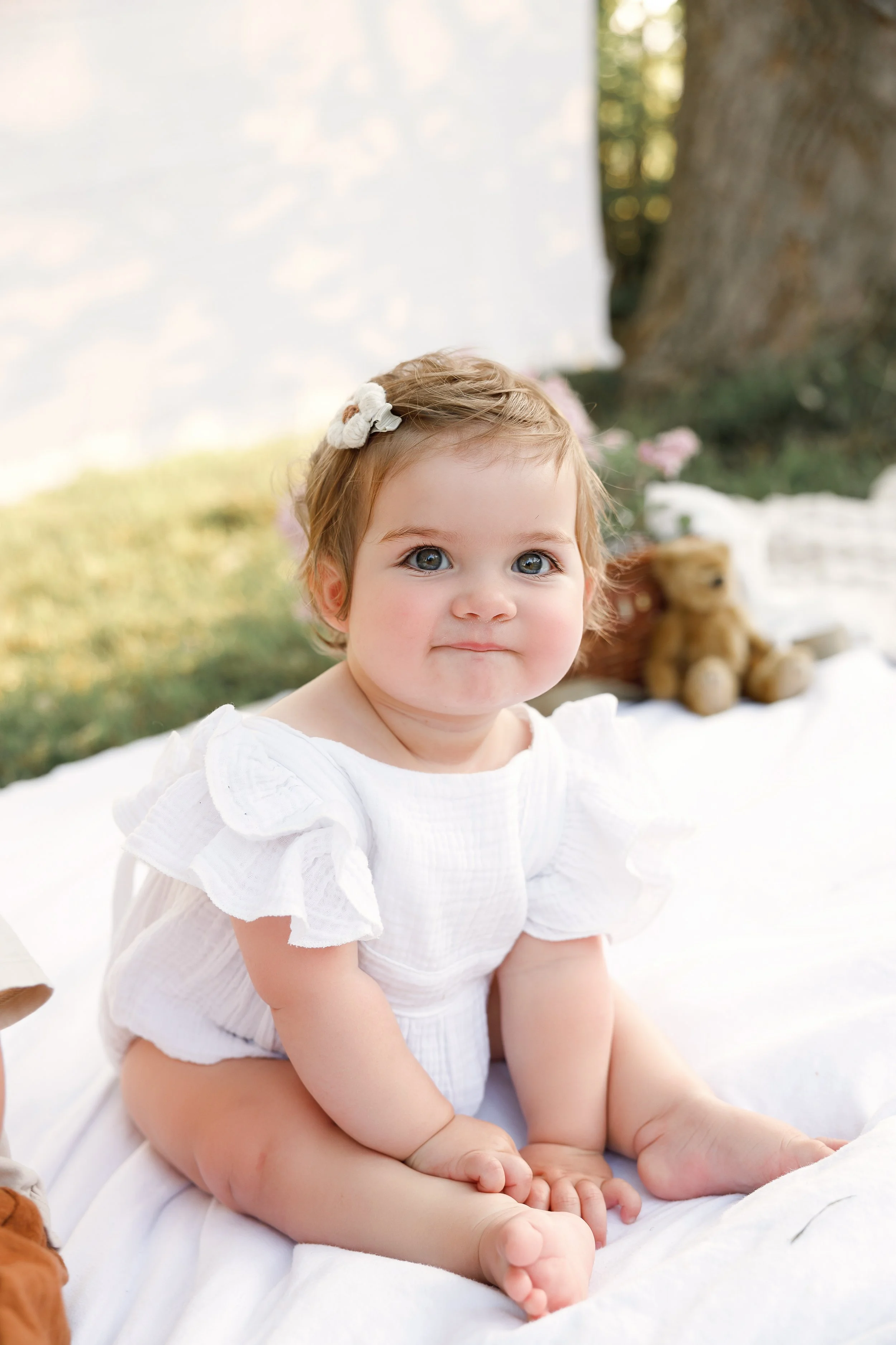 Cute baby girl with blue eyes and a hair bow, wearing a white dress with ruffled sleeves, sitting on a white blanket outdoors near a tree with a teddy bear in the background.