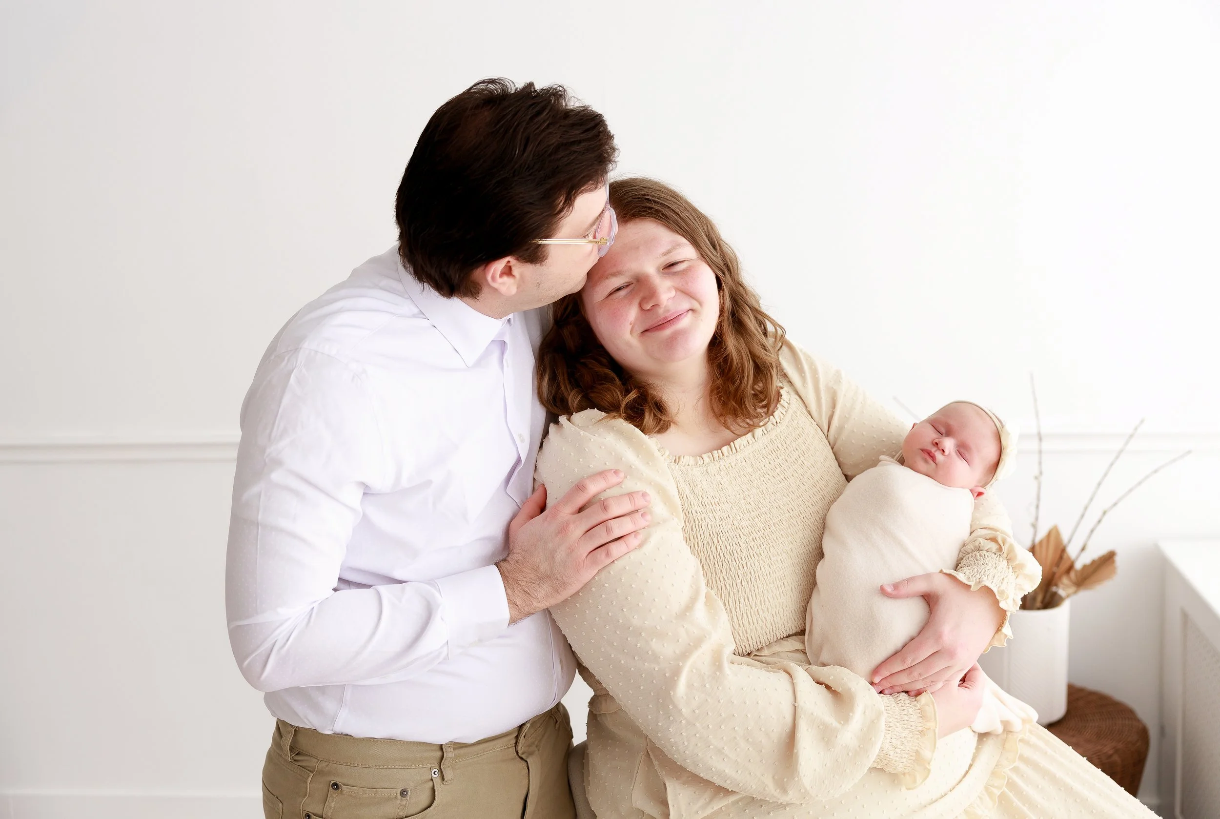 A young girl with curly hair being held by a woman, who is holding a newborn baby, in a bright room with white walls and a potted plant in the background.
