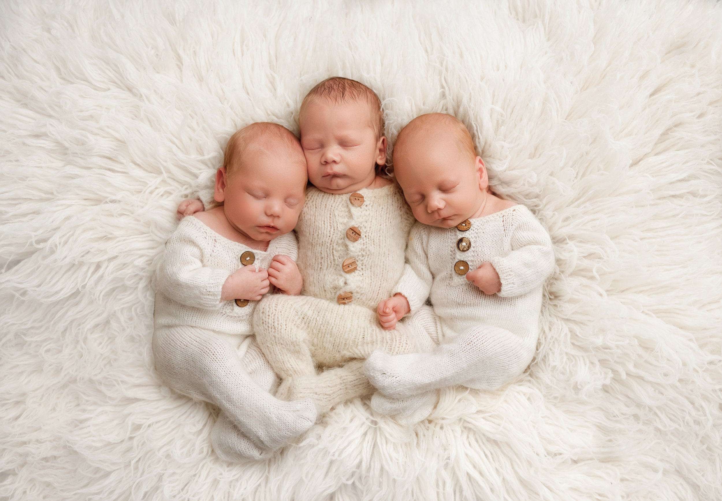 Three newborn babies sleeping on a white fluffy blanket, dressed in white knitted outfits with wooden buttons.