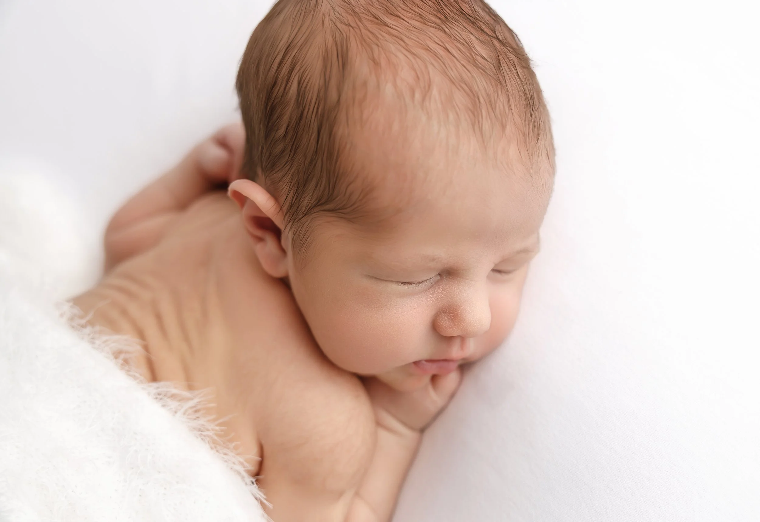 Close-up of a sleeping newborn baby with light skin and fine hair, resting on a soft white surface.