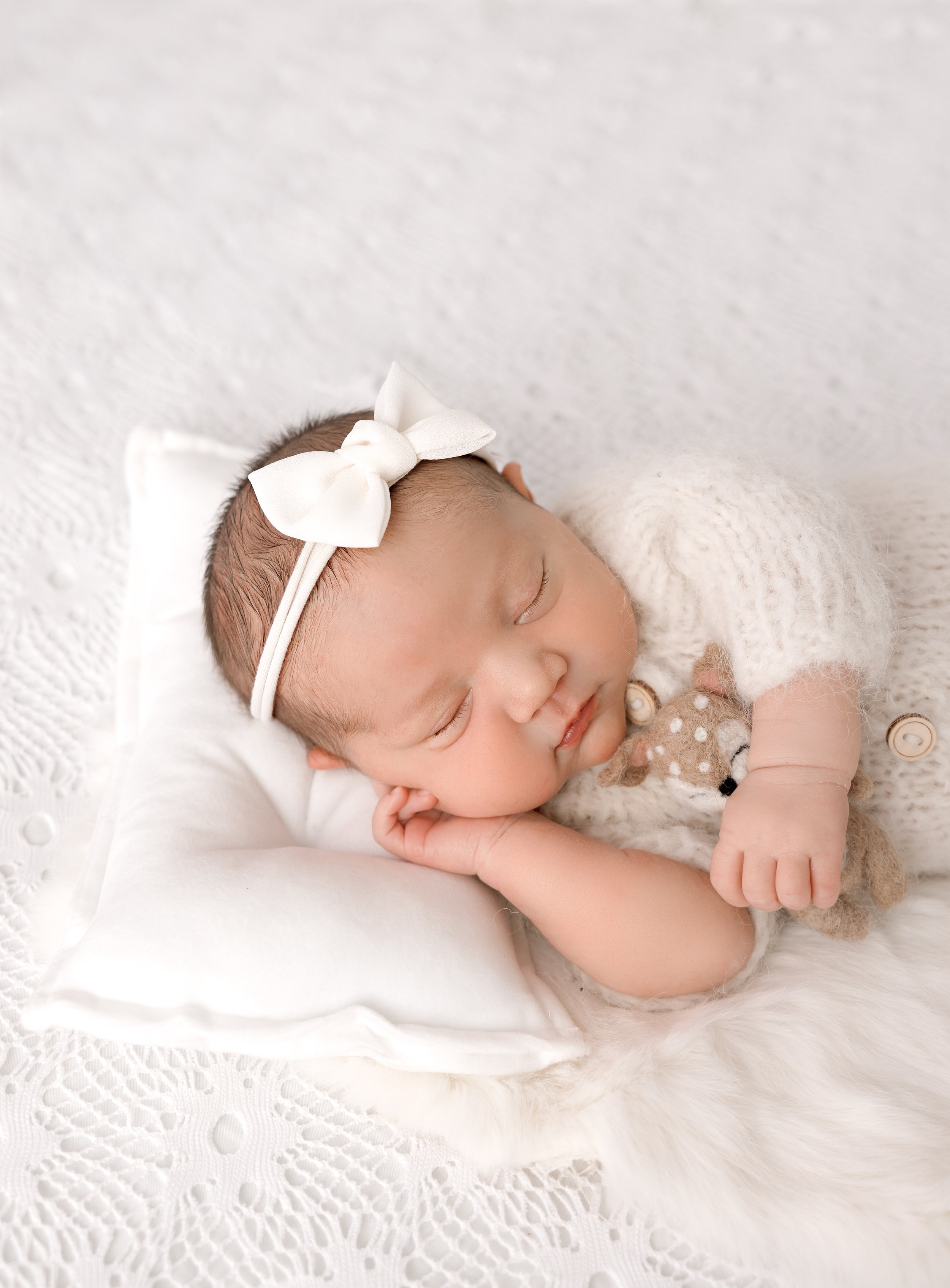 Close-up of a sleeping newborn baby girl with a white bow headband, resting on a white textured blanket with a stuffed animal nearby.