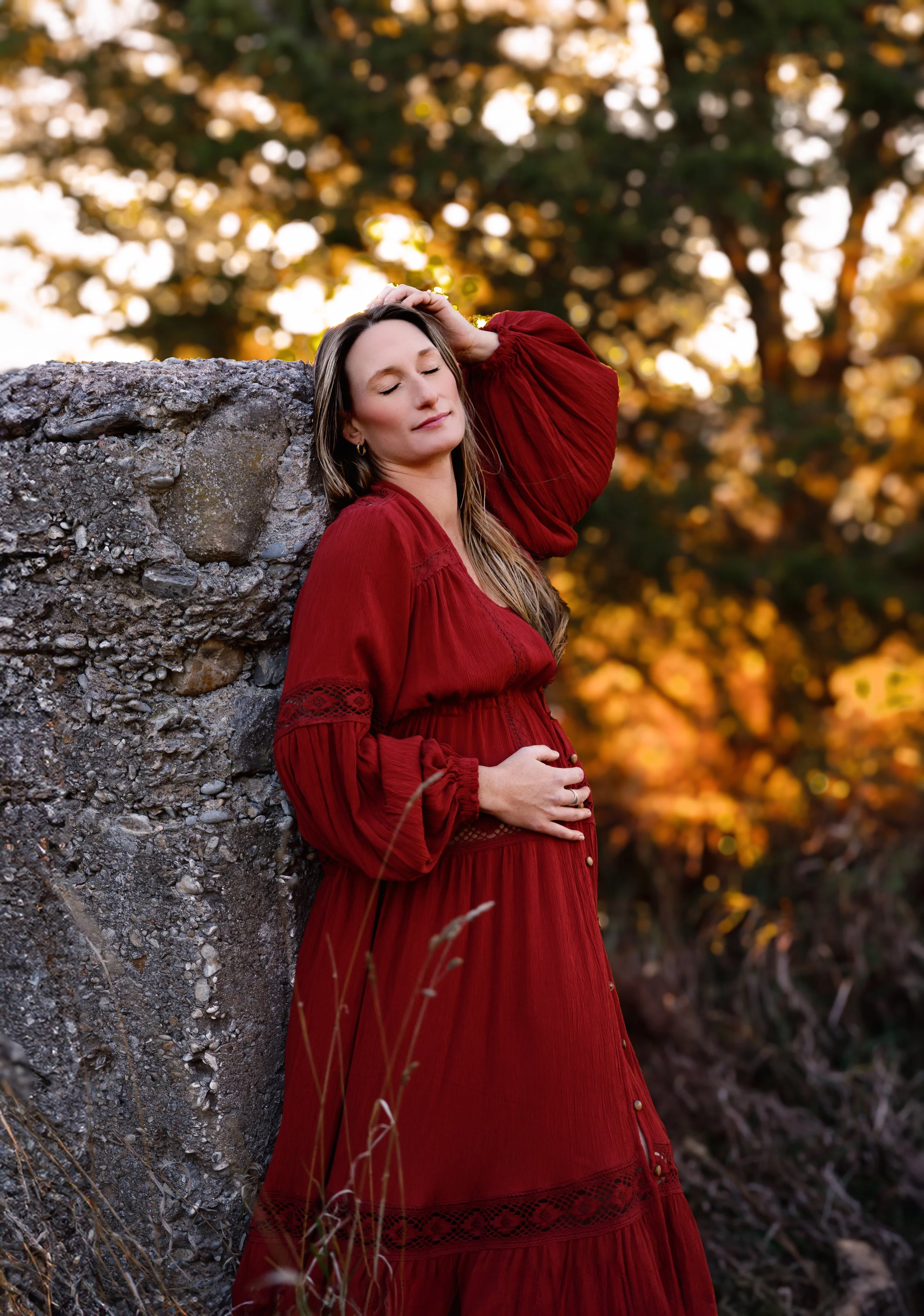 A woman in a flowing red dress with long hair, standing outdoors near a large stone, with her eyes closed and one hand touching her head, during autumn with golden leaves in the background.