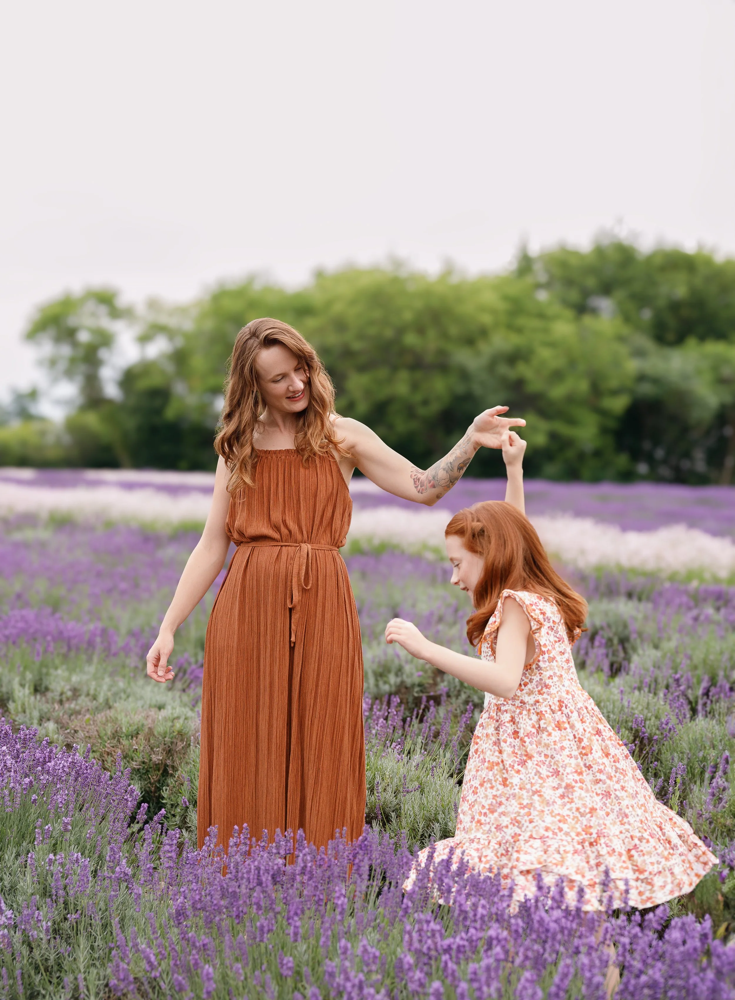 A woman and a young girl with red hair and matching floral dresses in a lavender field. The woman is smiling and pointing, while the girl is looking at her, crouching in the flowers.