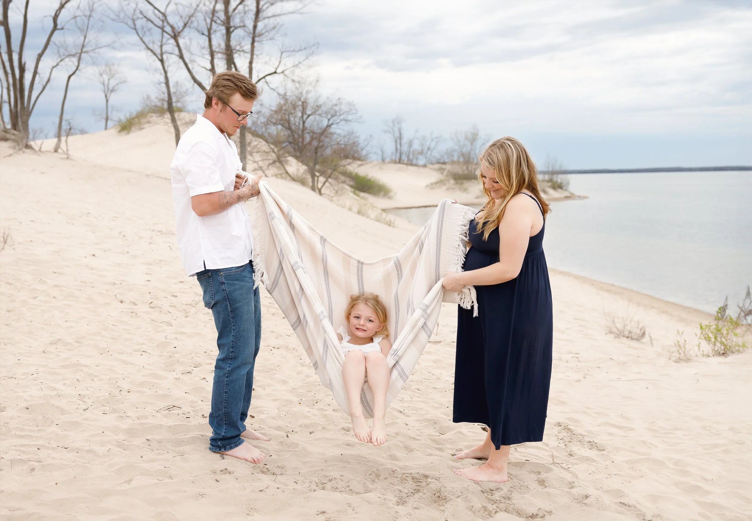 A family at the beach, with two adults and a young girl in a hammock. The man has glasses and tattoos, the woman is smiling, and the girl is sitting with her feet up, all standing on sand near the water with trees in the background.