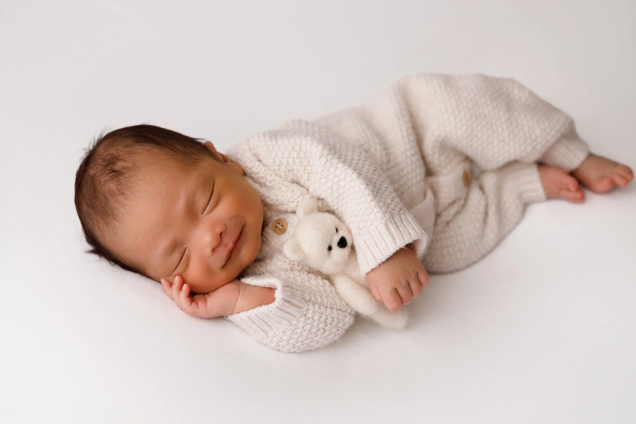 A sleeping infant lying on a white surface, wearing a knitted cream-colored outfit with bear buttons and holding a small white stuffed bear.