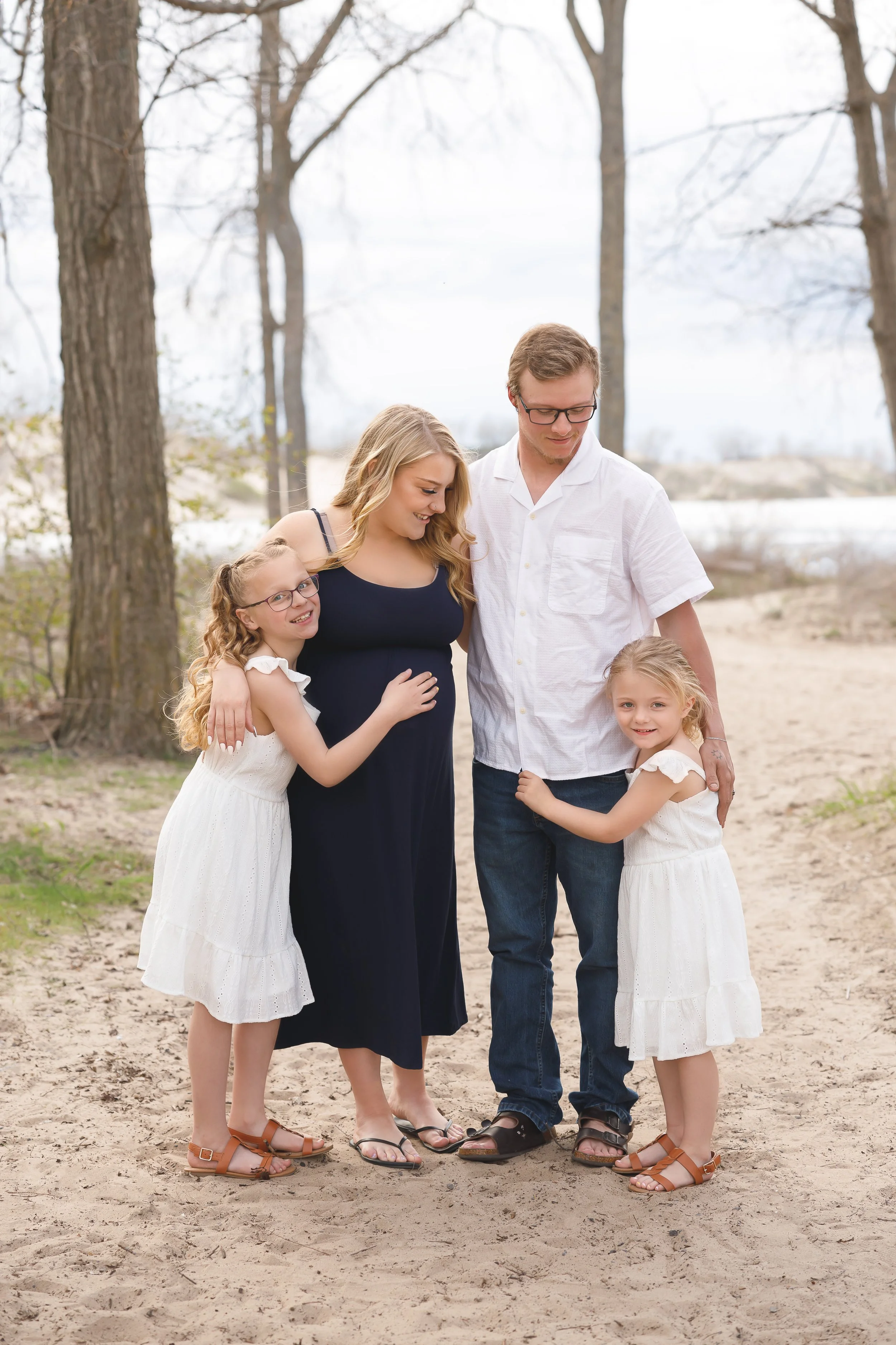 A family of five standing close together outdoors on a sandy area, surrounded by leafless trees, with a body of water in the distance. The family includes two young girls and a man and woman who appear to be their parents.
