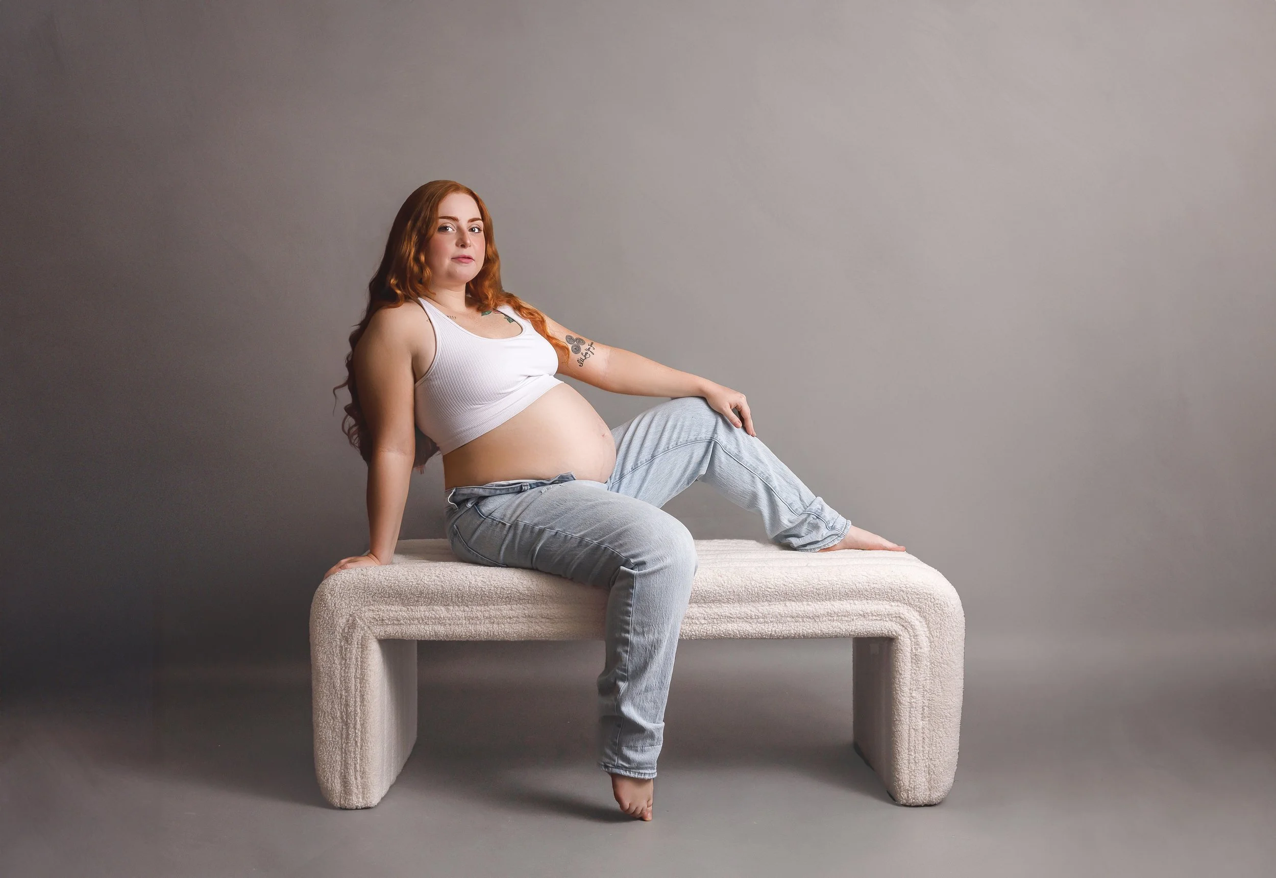 A pregnant woman with long red hair is sitting on a beige textured bench against a plain gray wall. She is wearing a white sleeveless top and light blue jeans, with her left arm resting on her knee and her right hand on the bench.