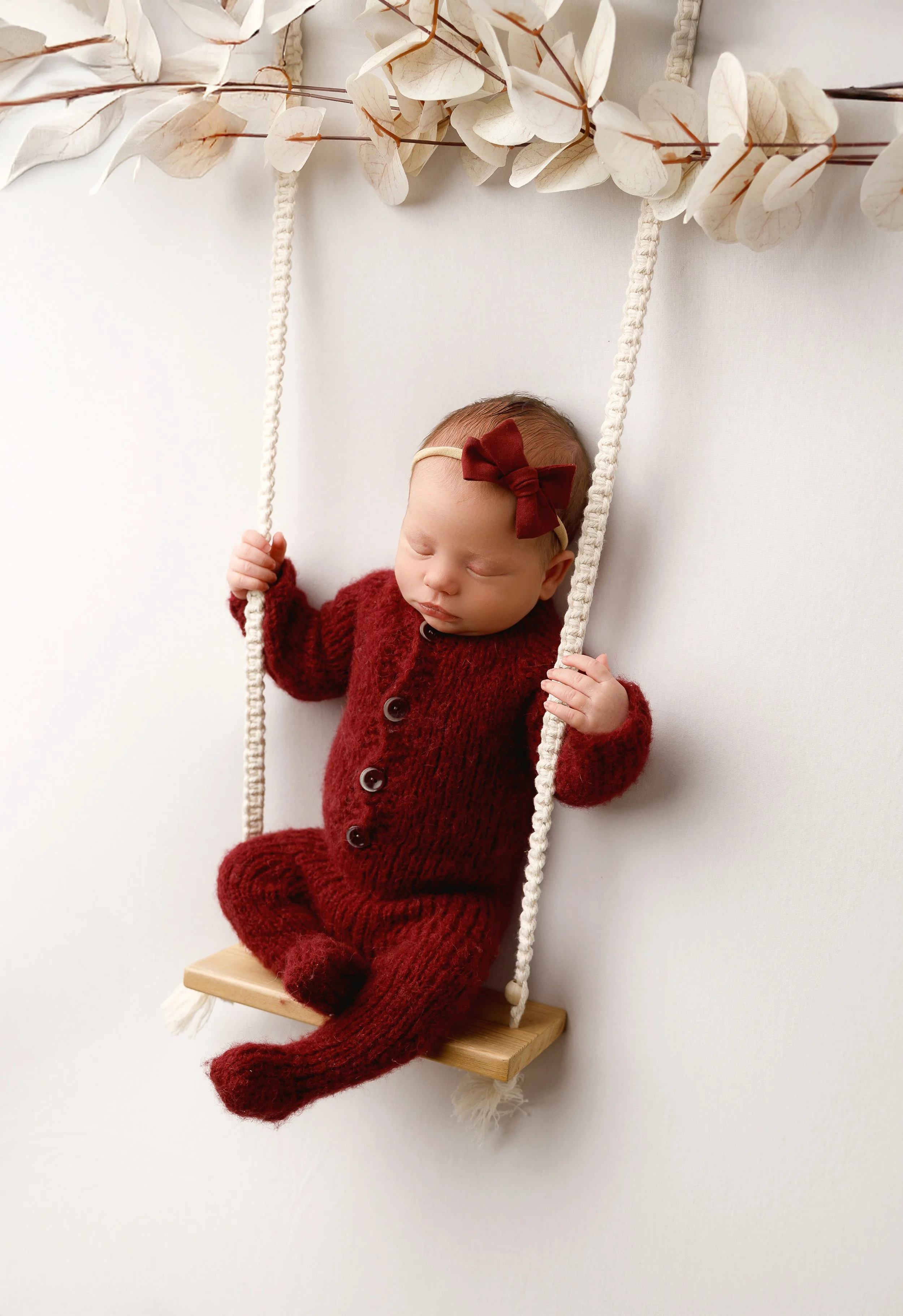 A sleeping baby dressed in a red knitted outfit, sitting on a small wooden swing with white ropes, against a plain white background. The baby is wearing a cream-colored headband with a red bow.
