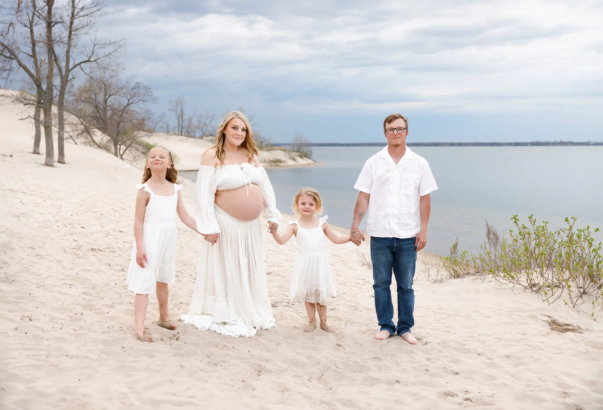 A family of five standing on a sandy beach near water, dressed in white, with three young girls, a pregnant woman, and a man with tattoos, under a cloudy sky.