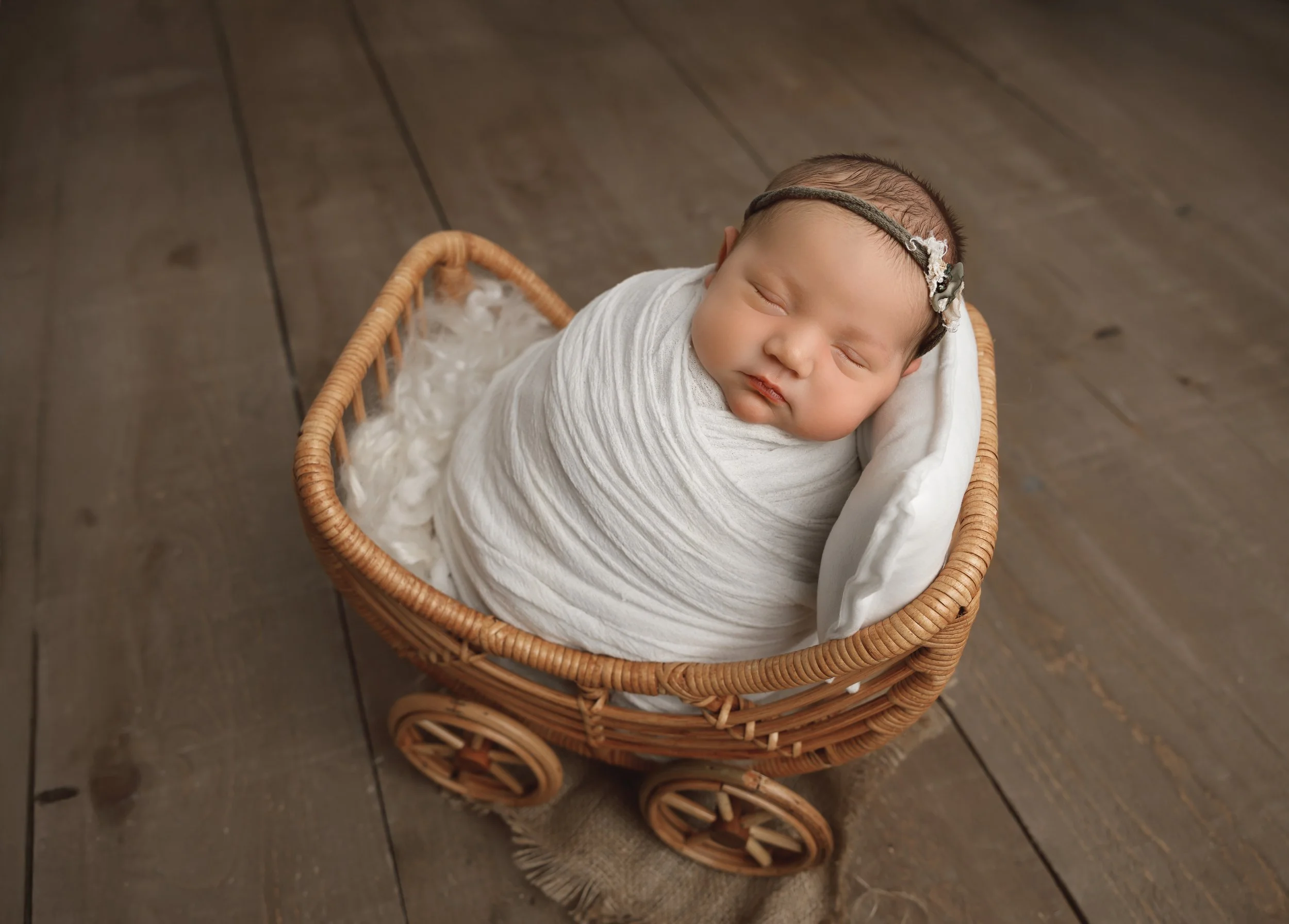 A sleeping baby wrapped in light-colored swaddle blankets, lying in a small wicker toy stroller with wheels on a hardwood floor.