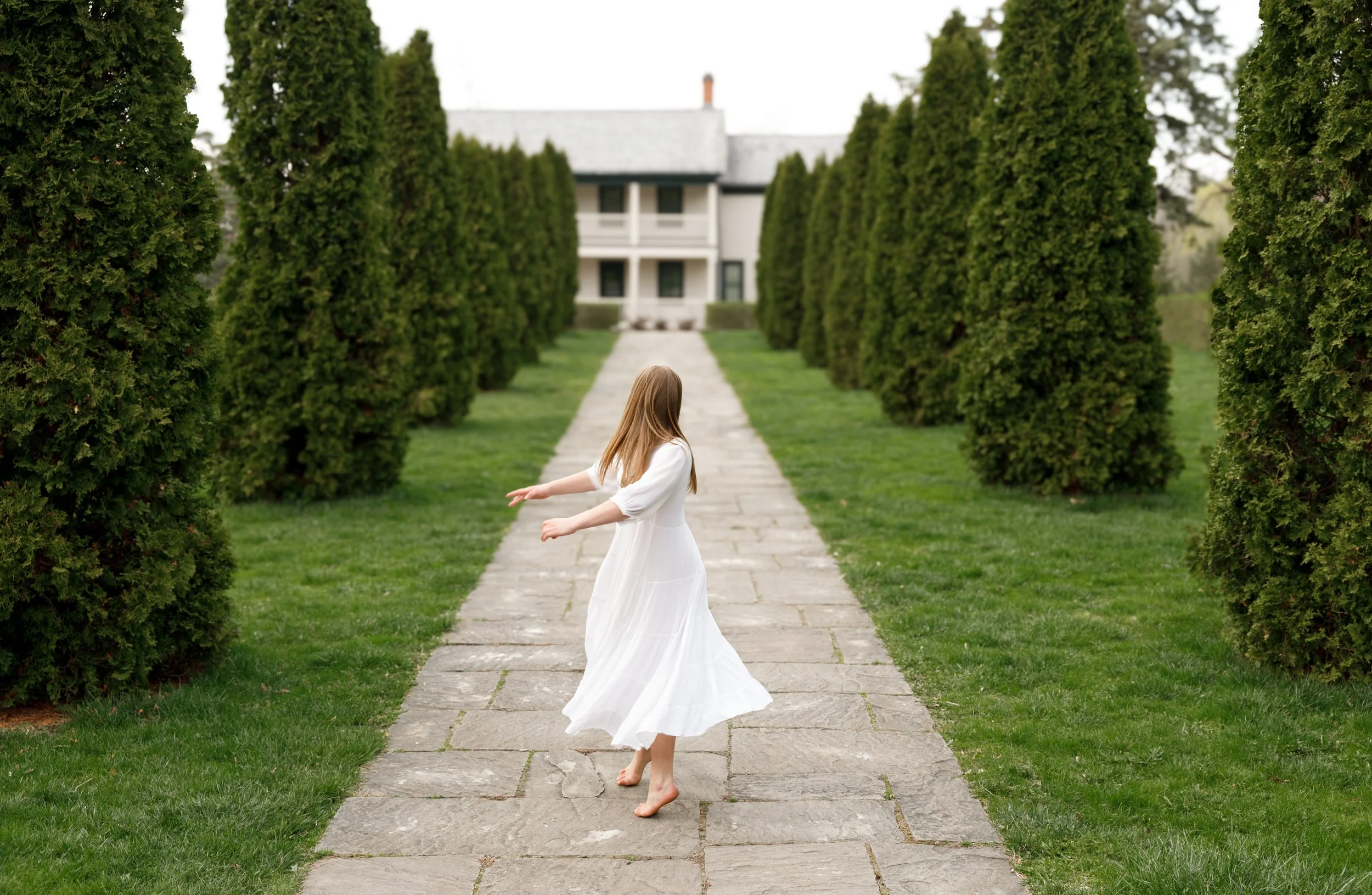 A young girl in a white dress is spinning around barefoot on a stone pathway lined with tall green bushes leading to a white house with black windows and a porch, in a lush green garden.