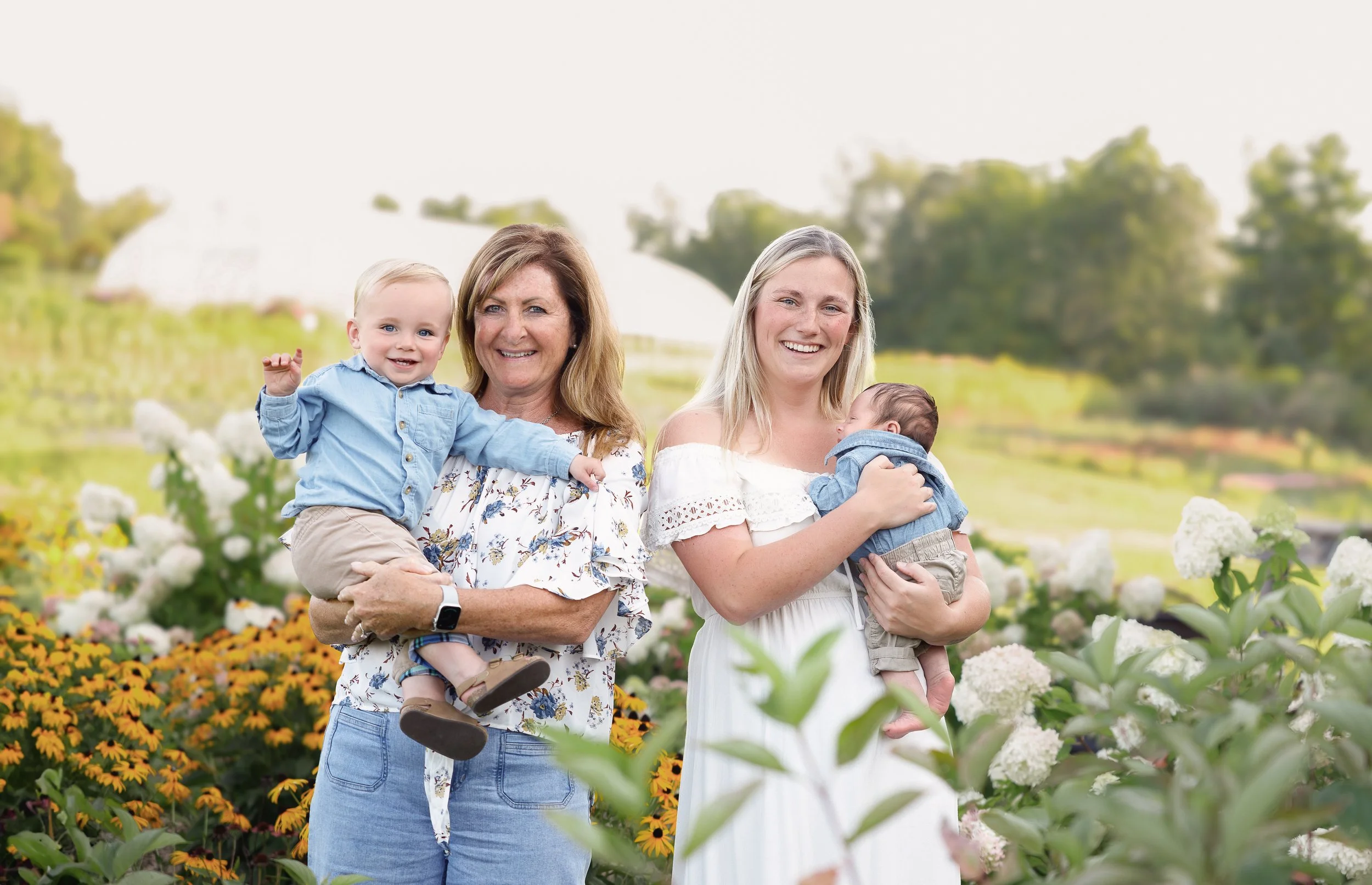 Family of four smiling outdoors among flowers, with a woman holding a baby and an older woman holding a young boy, in a green and white landscape.