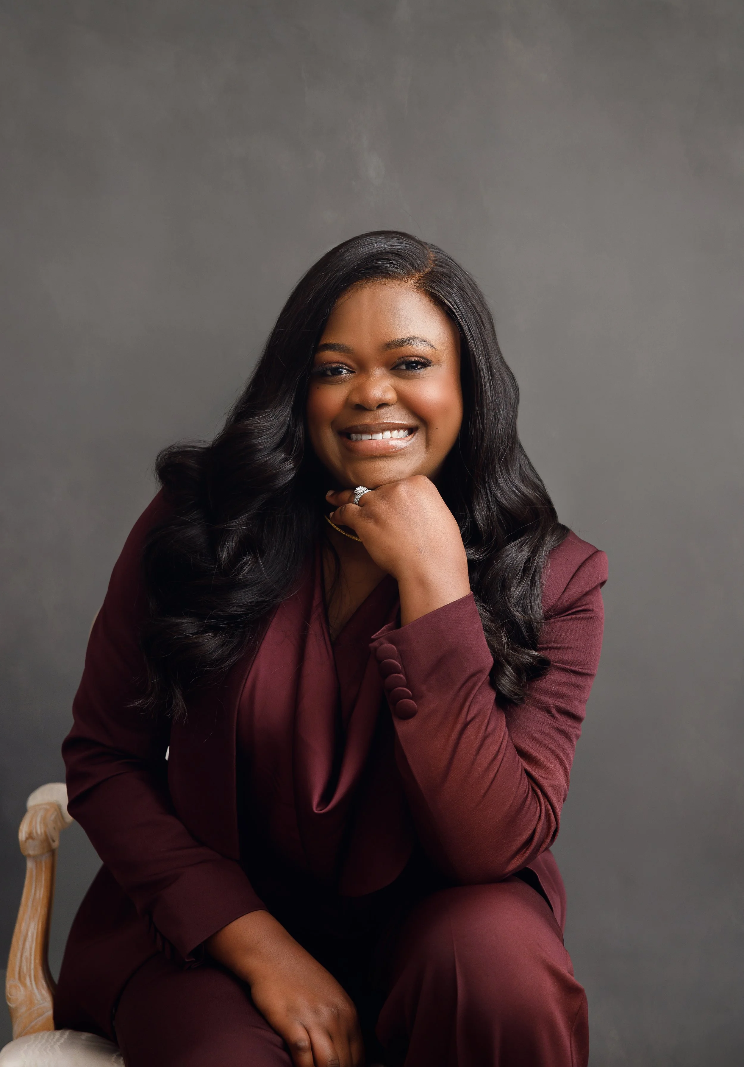A woman with long black hair wearing a burgundy suit, sitting on a vintage wooden chair, smiling with her hand resting on her chin, against a plain gray background.