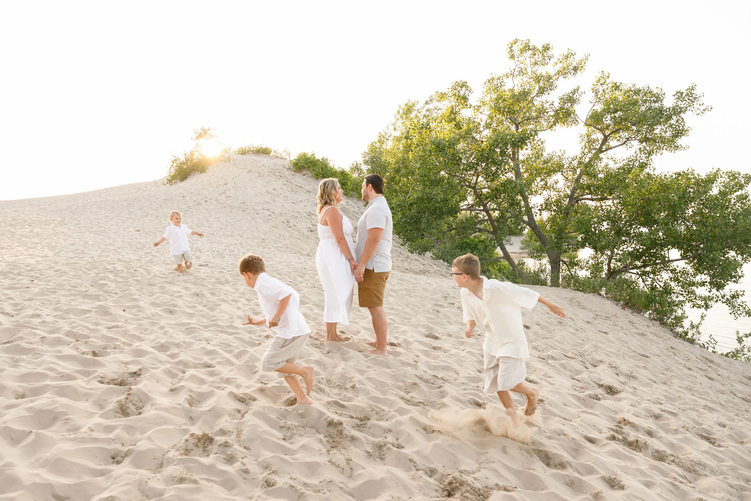 A couple standing hand in hand on a sandy dune with children playing around them, and trees in the background, during sunset.