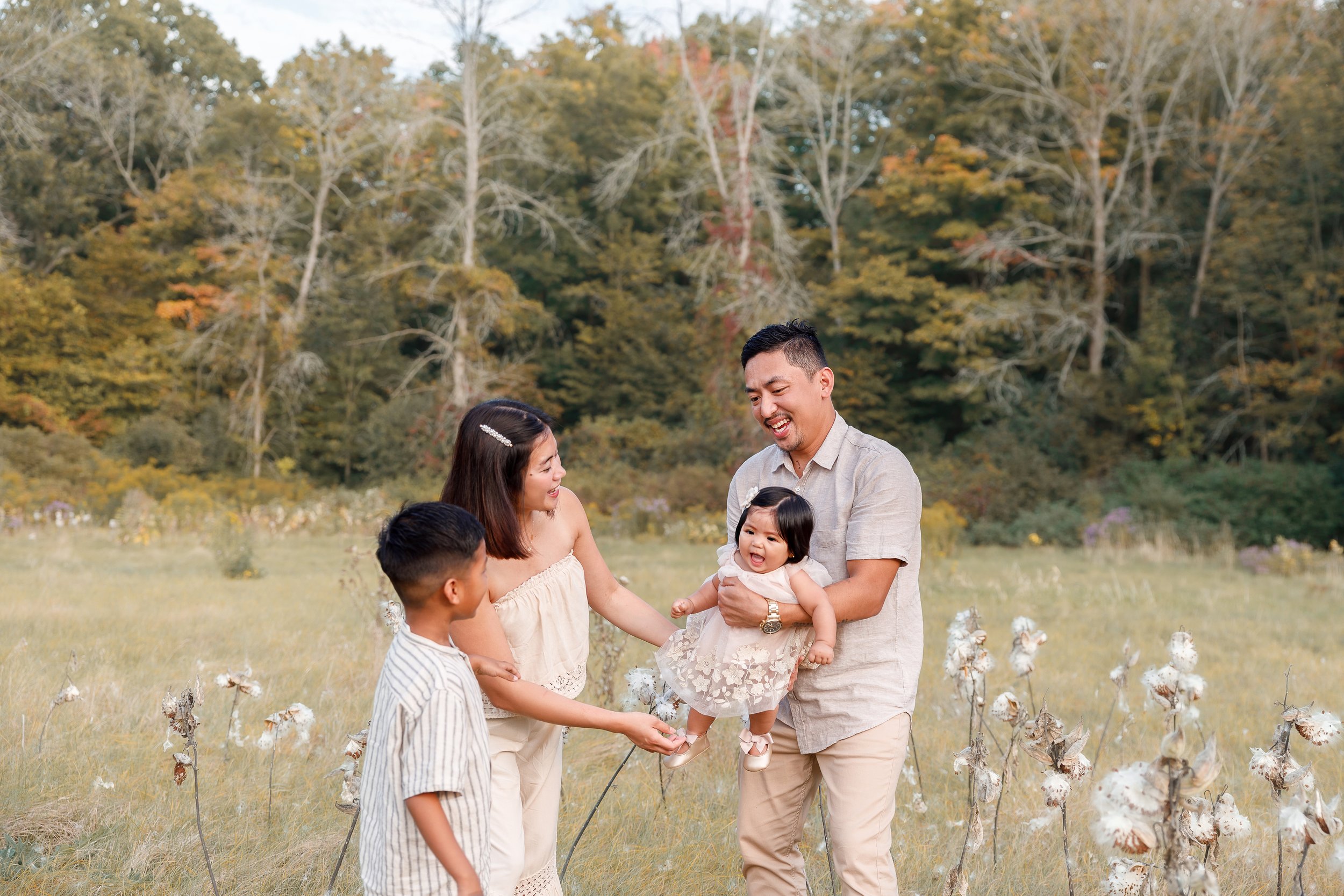 A family enjoying an outdoor fall day, with a man holding a smiling young girl in a cotton dress, standing in a cotton field surrounded by cotton plants and trees with autumn-colored leaves.