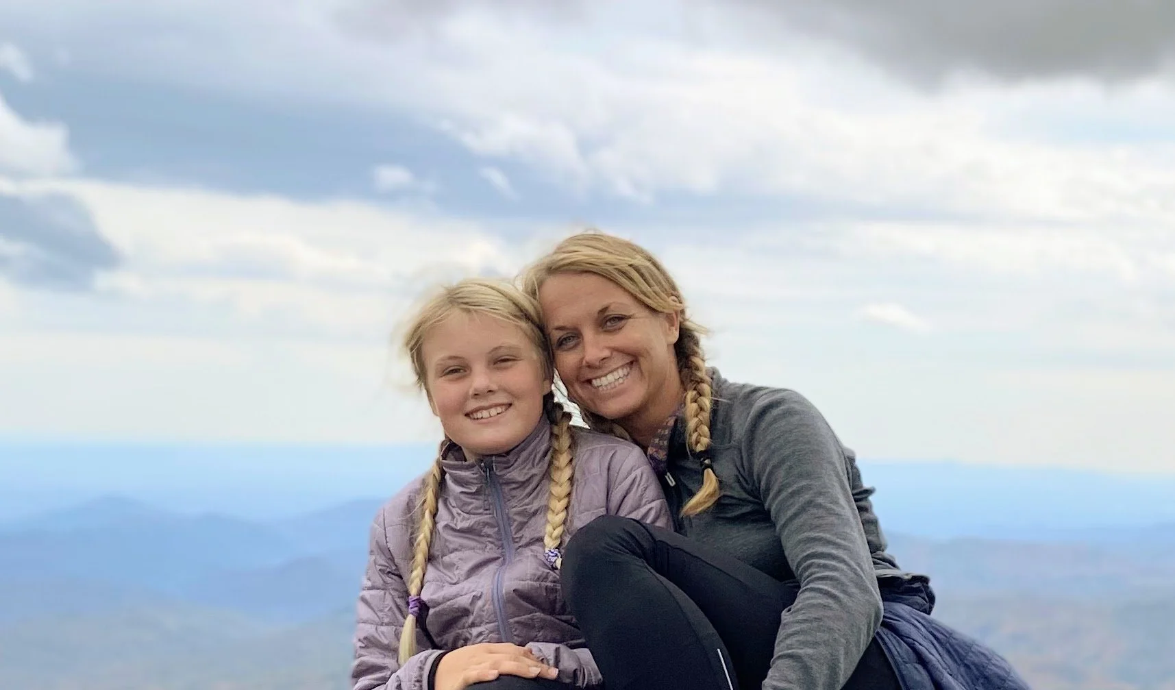 A woman and a young girl with blonde hair and braided pigtails smiling and sitting close together outdoors against a cloudy sky and mountain landscape.