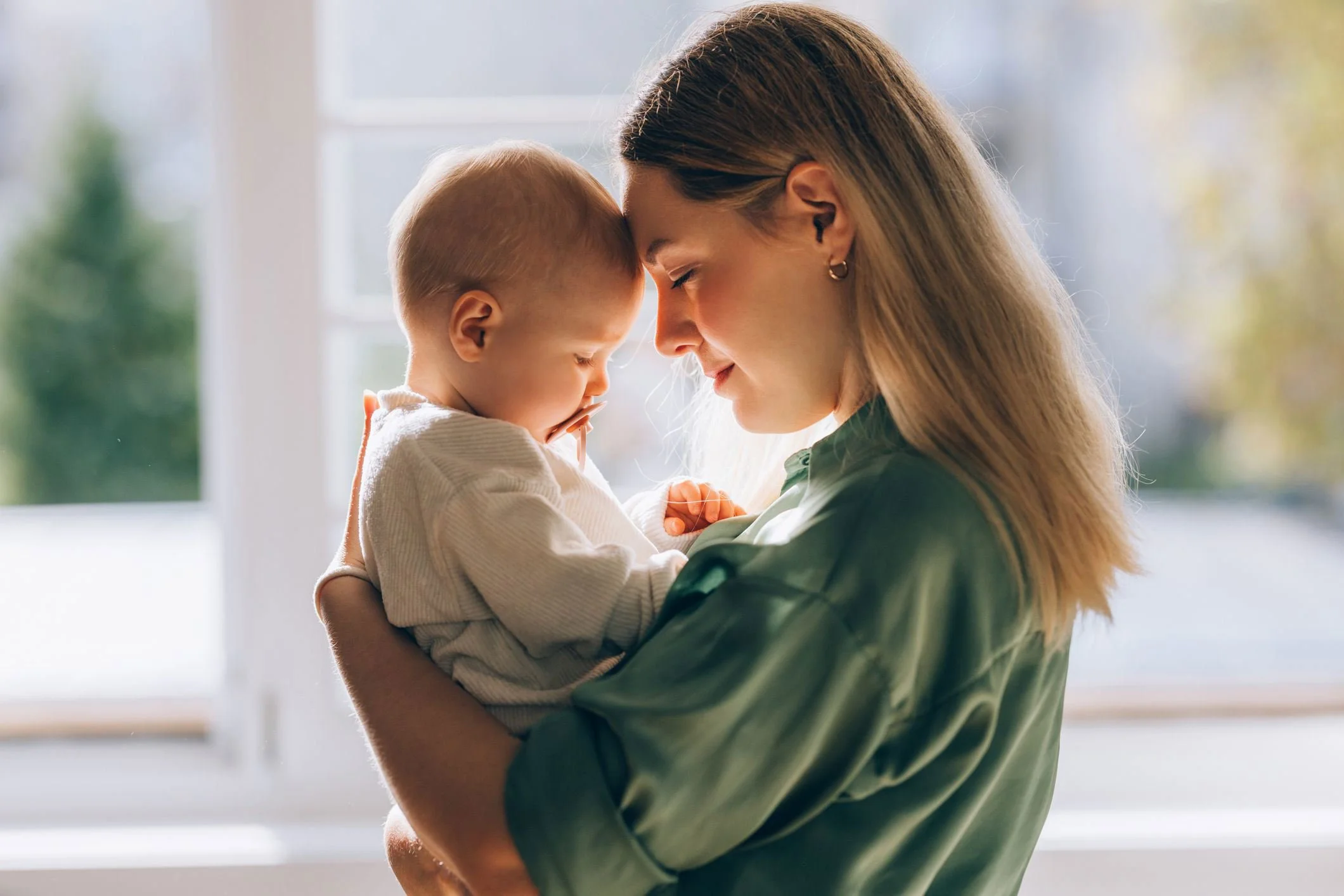 A woman holding a young child close, both touching foreheads with gentle smiles inside a bright room with large windows and blurred outdoor scenery.