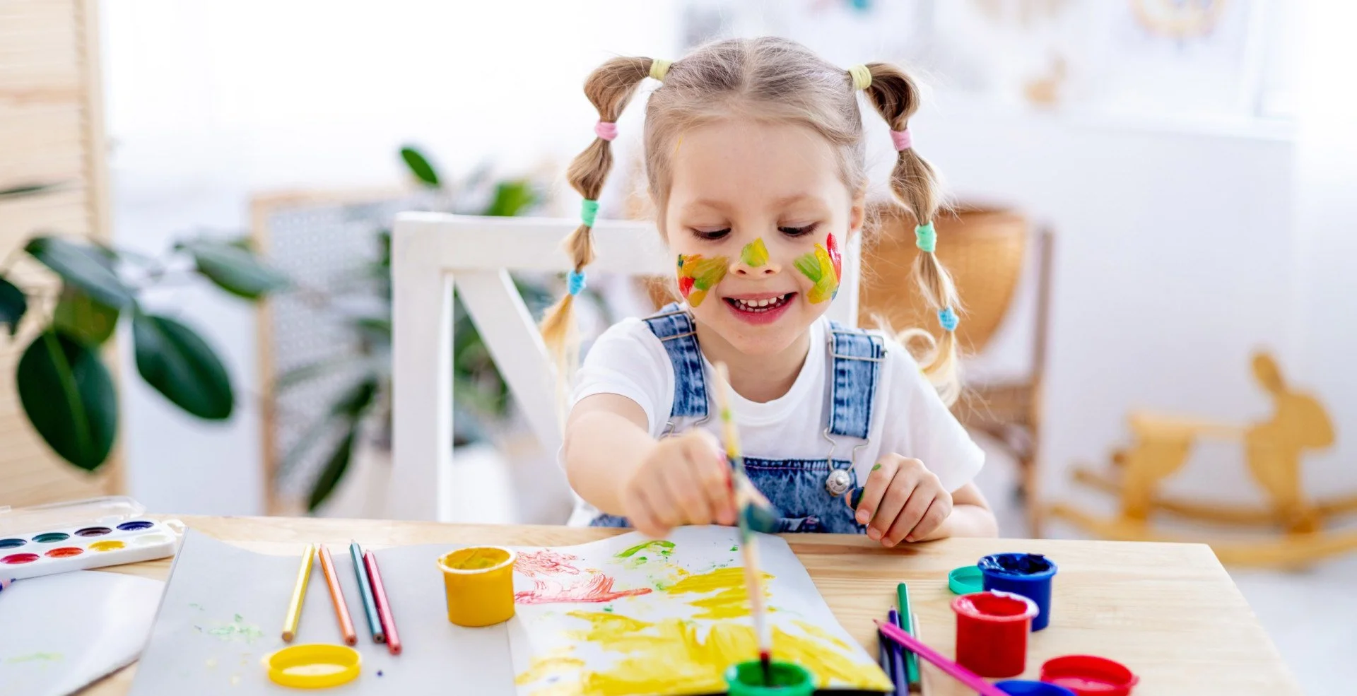 A young girl with pigtails and colorful face paint is painting with watercolors at a table, smiling and enjoying art.