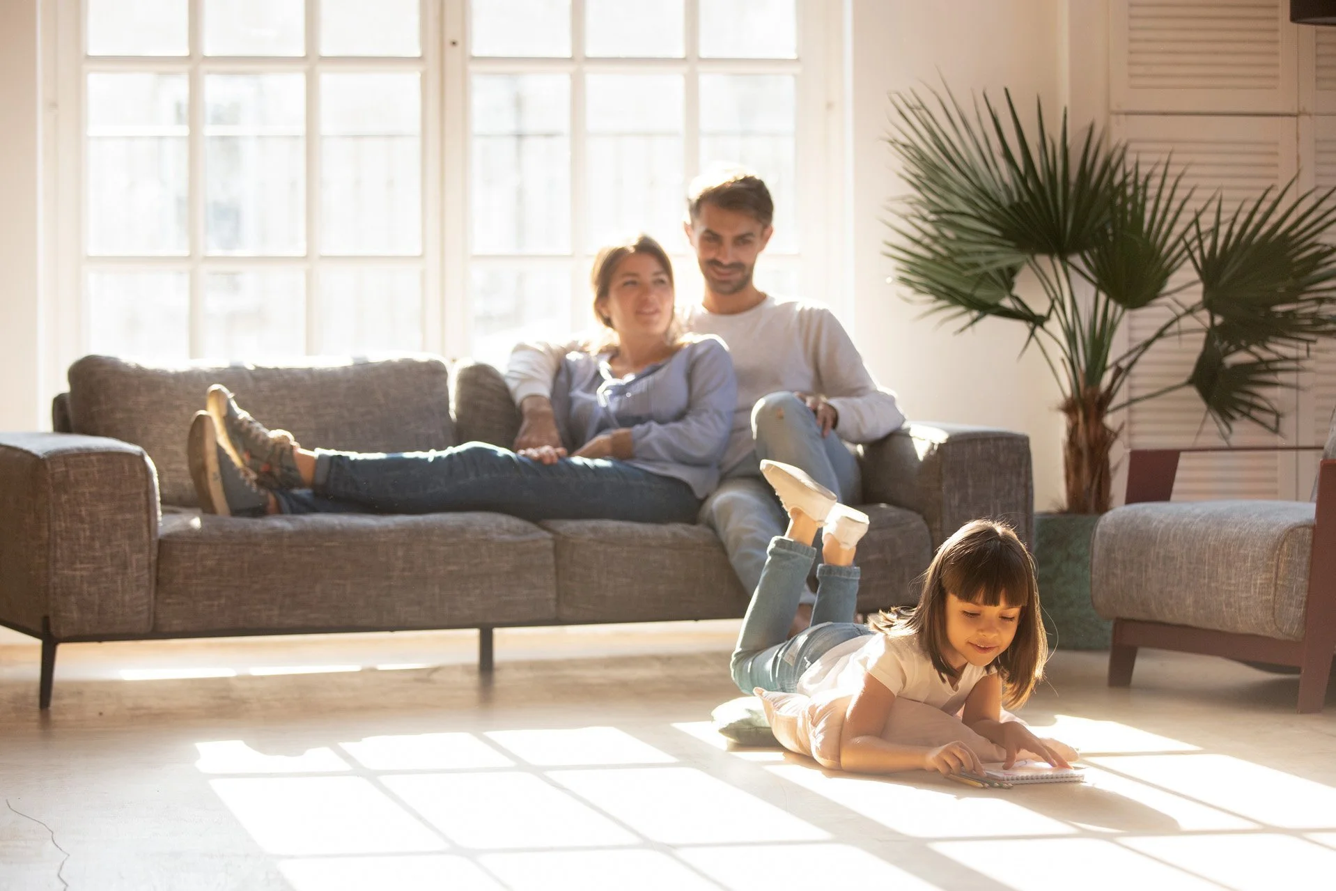 A man and woman sitting on a gray couch in a sunlit living room, with a young girl lying on the floor playing with a book in the foreground, and another child lying on the floor in front of the girl with their legs raised.