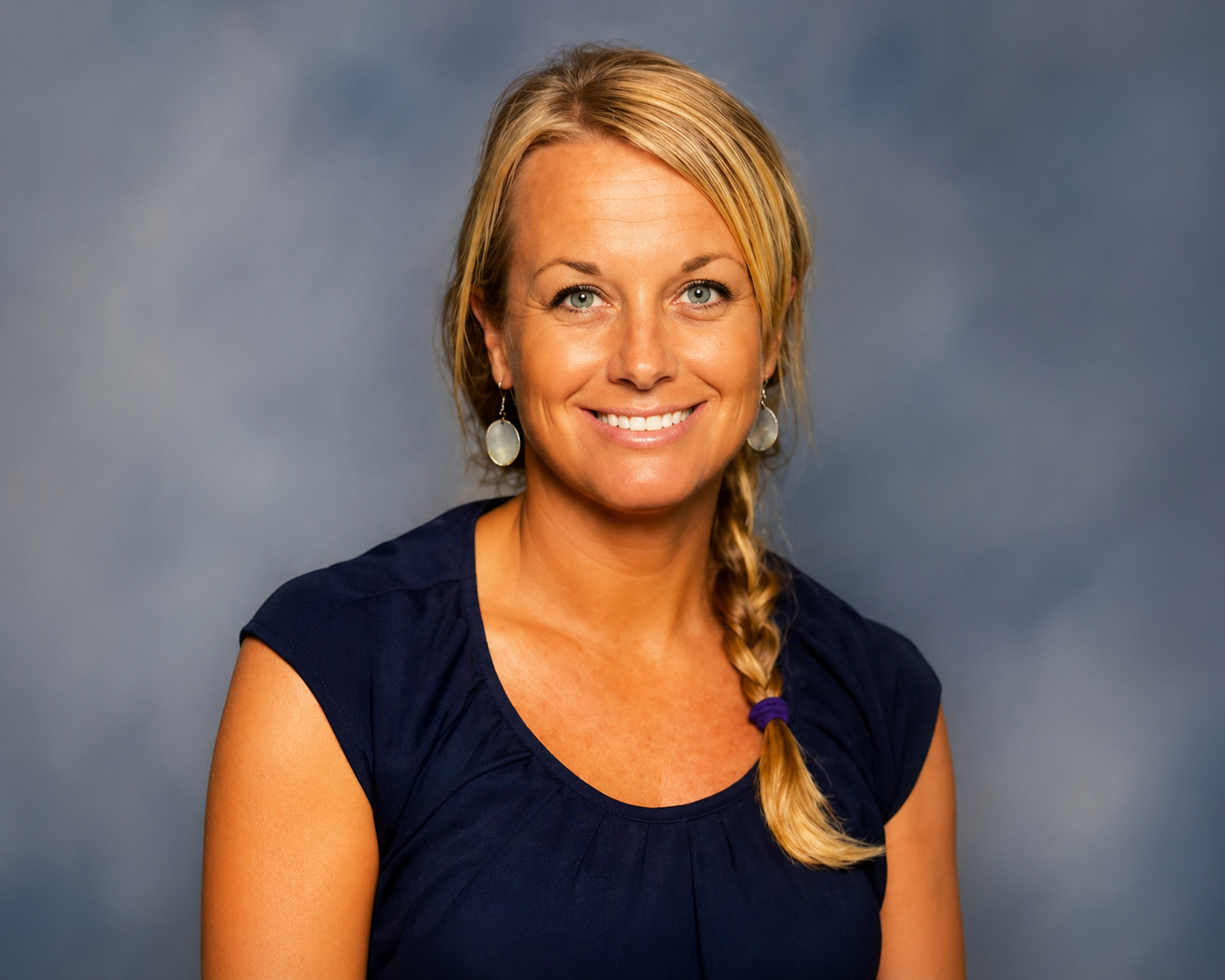 A smiling woman with blonde hair in a side braid, wearing a navy blue top and large white earrings, against a plain background.