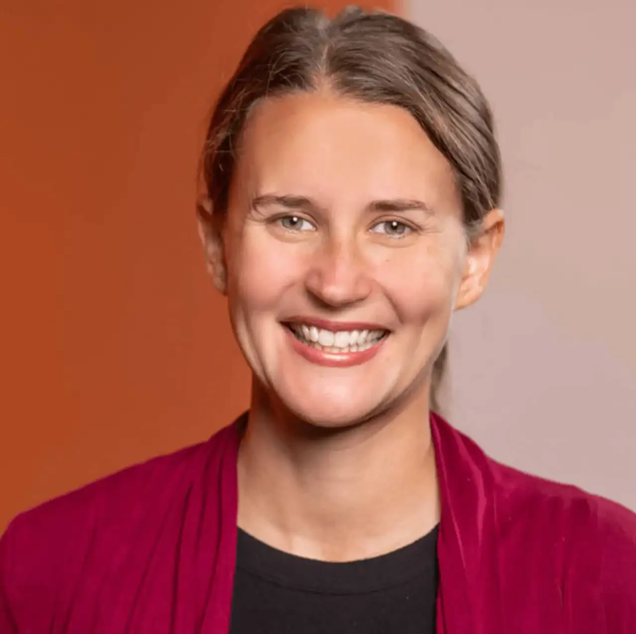 Woman smiling and wearing a pink sweater with a black shirt and a orange and pink background.