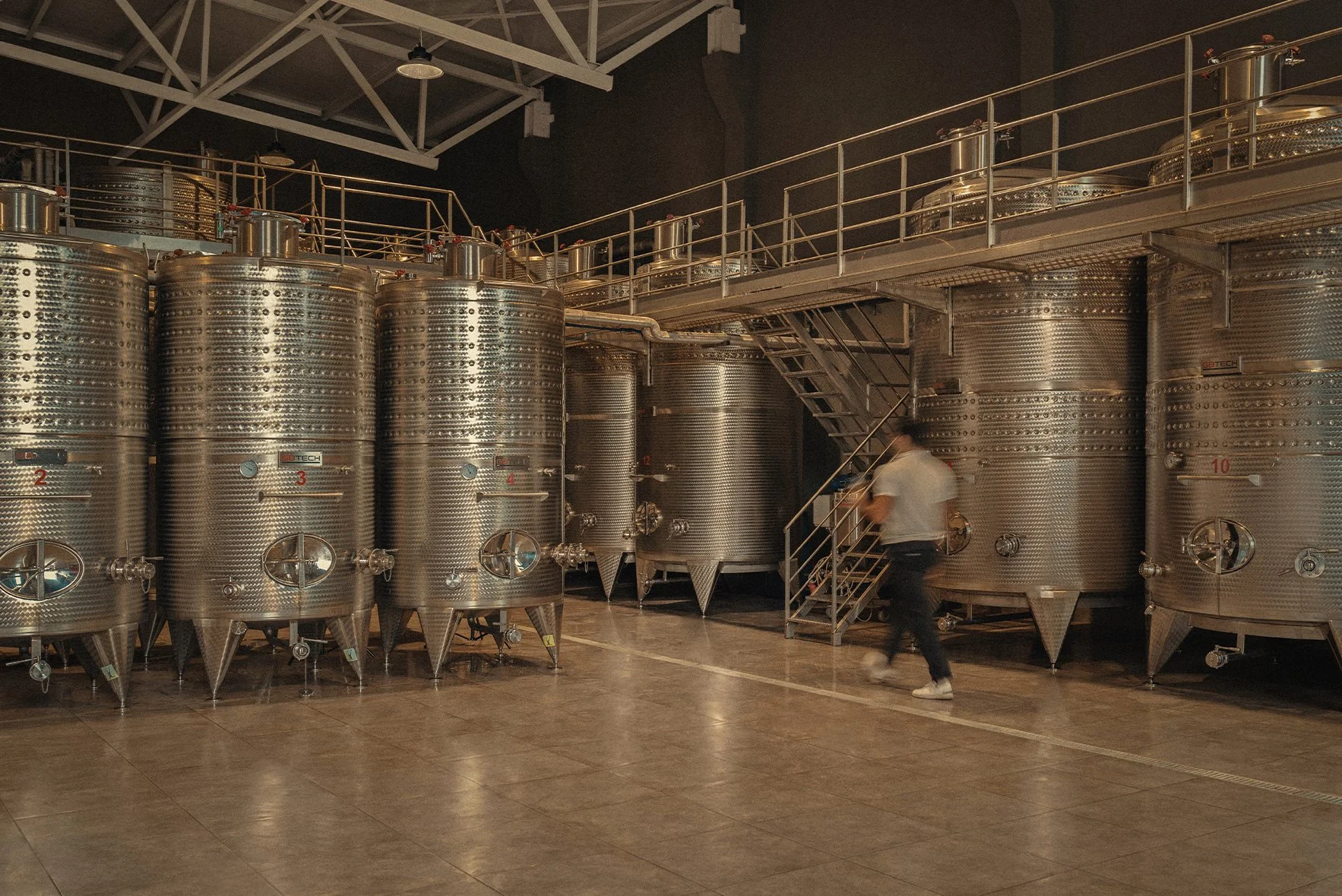 A large industrial winery or brewery with multiple stainless steel fermentation tanks and a person walking on a staircase pathway.