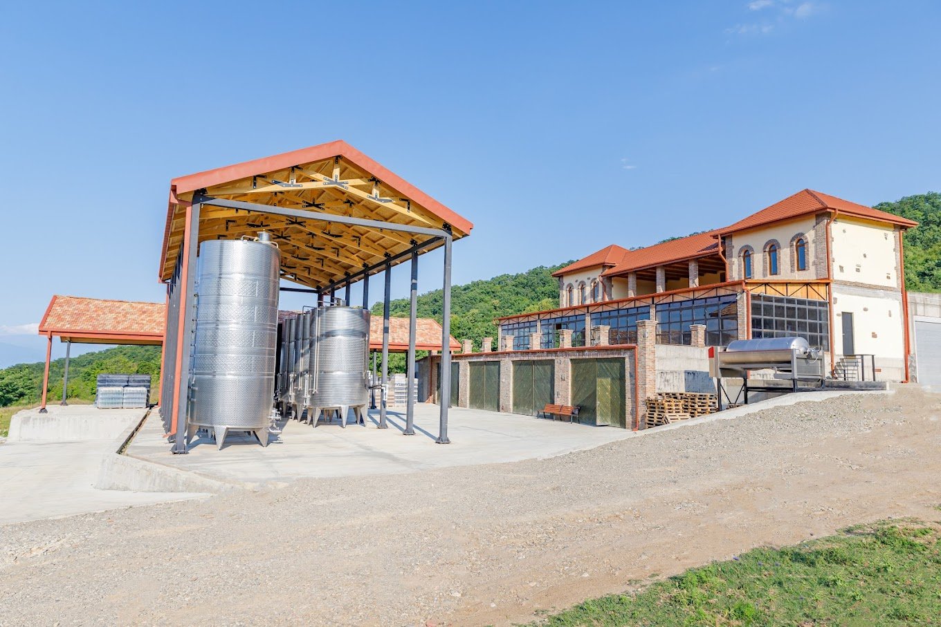 View of a winery with large stainless steel fermentation tanks under a wooden roof structure, alongside a building with a red tile roof and a glass-enclosed patio, set against green hills and a clear blue sky.