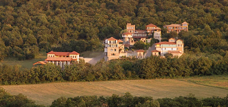 A large estate or mansion complex situated on a hillside, surrounded by dense green trees, with multiple buildings featuring red-tiled roofs and stone walls.
