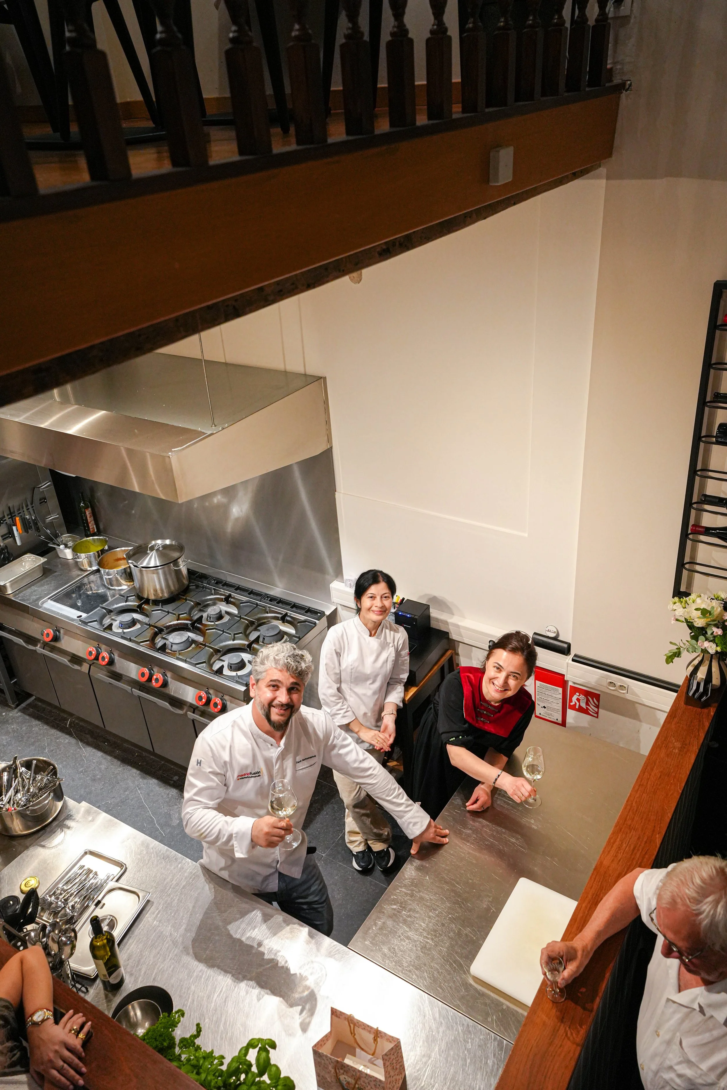 A group of smiling people in a kitchen, some holding glasses of white wine, with a chef in a white uniform in the foreground, chefs and kitchen equipment visible behind them.