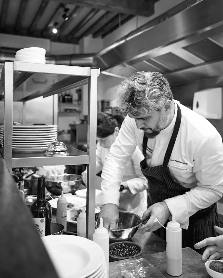 Chef in a busy kitchen preparing food, with shelves of plates and cooking supplies around him.