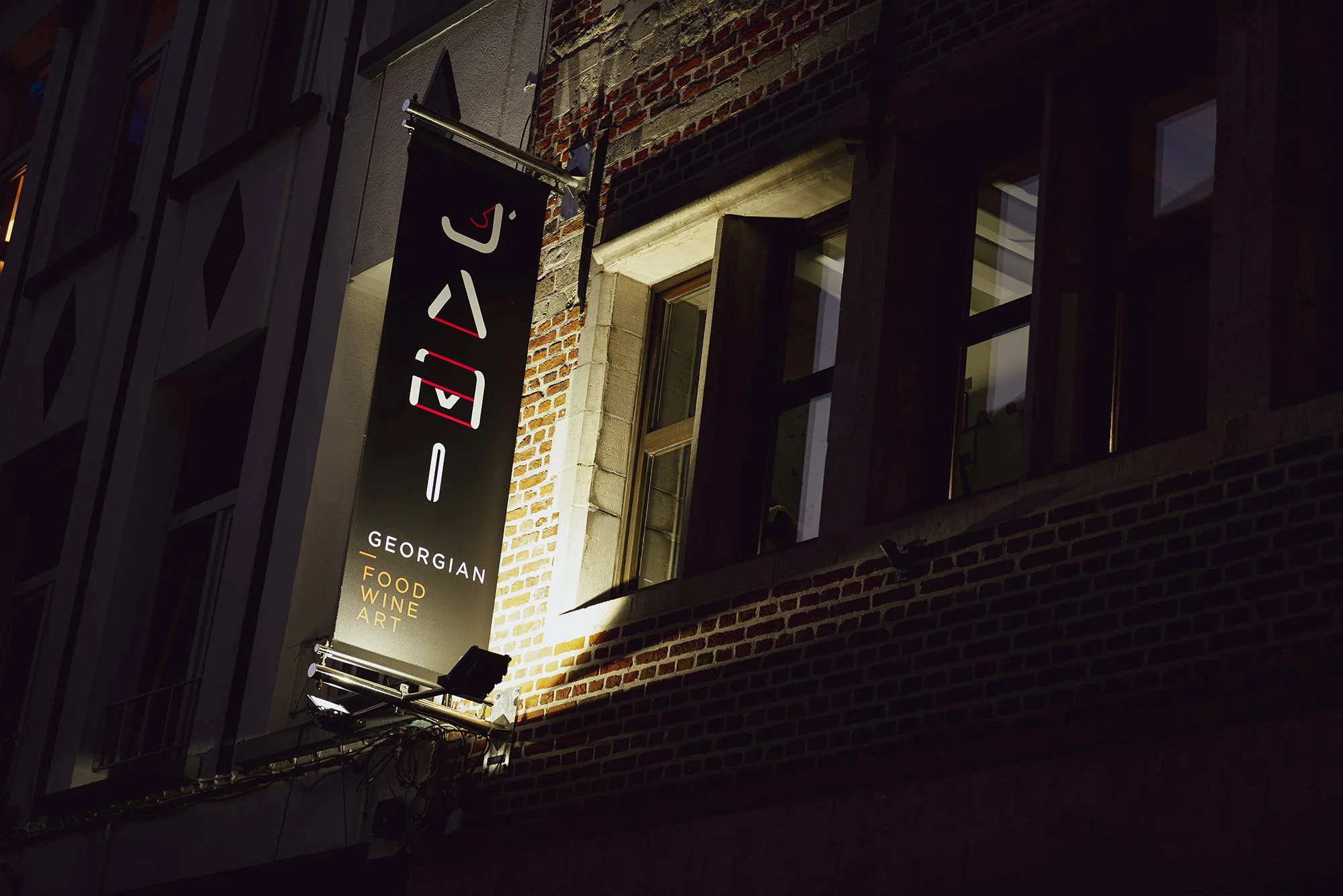 Nighttime view of a vertical black and white sign on a brick building that reads 'I A L E' with an arrow pointing left. Text on the sign below states 'GEORGIAN FOOD WINE ART'. The building's windows are visible, illuminated from inside, with some open shutters.